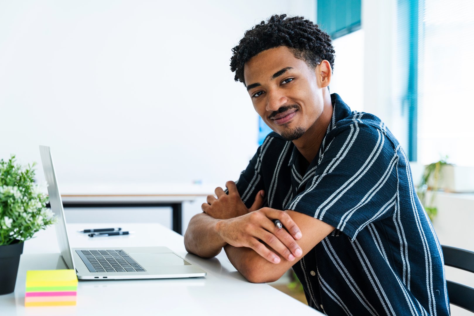 Young entrepreneur smiling and working using laptop in startup company office, modern workplace with plants and colorful sticky notes, successful businessman enjoying work