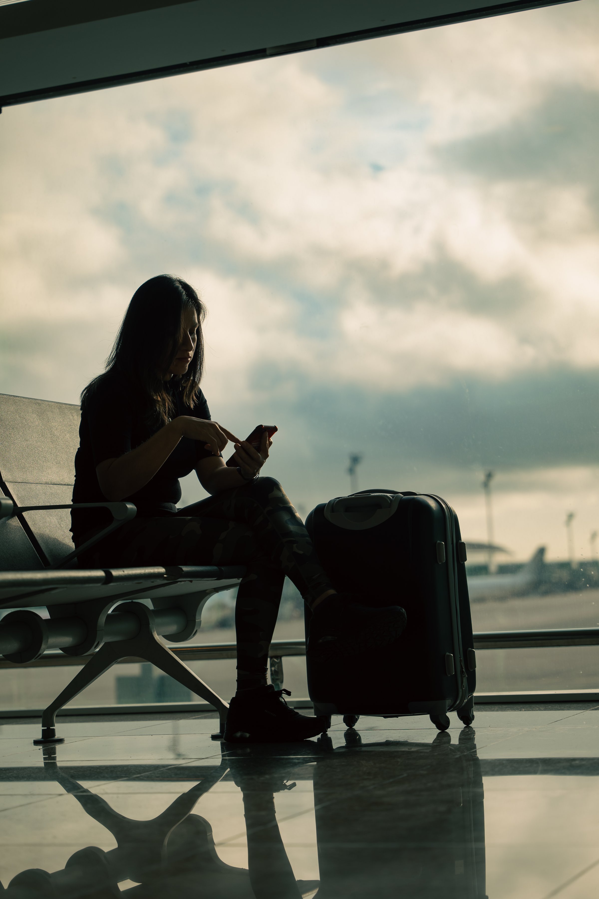 A woman with her suitcase checks her mobile phone at the airport while the cloudy sky is seen in the background.