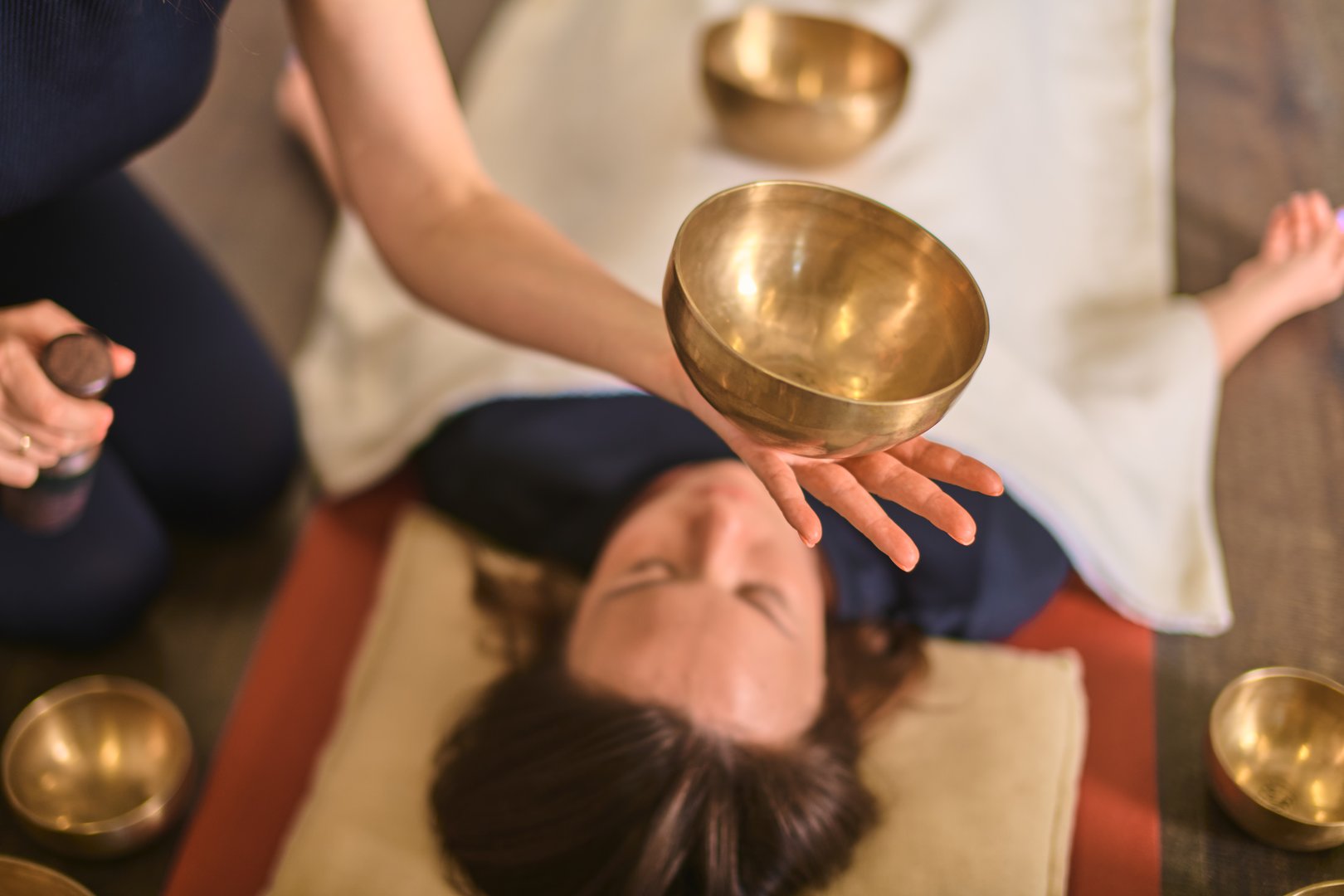 Young woman receives sound therapy with singing bowls in serene setting. Warm lighting enhances tranquil atmosphere. Hand gently holds bowl over her