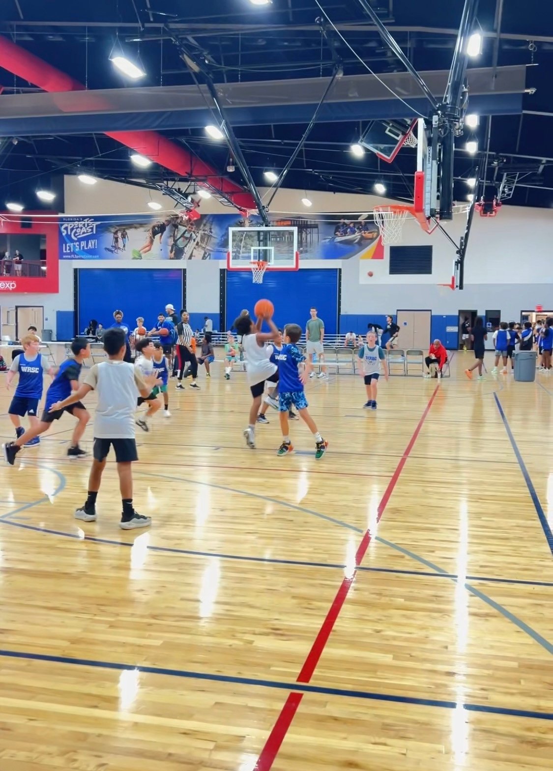Kids playing basketball in an indoor gym with a polished wooden floor and colorful walls.