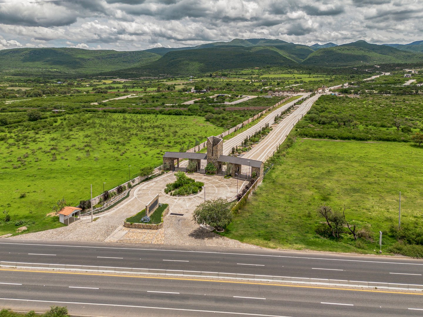 Aerial view of a gated entrance with roads and green fields in a mountainous landscape under cloudy skies.