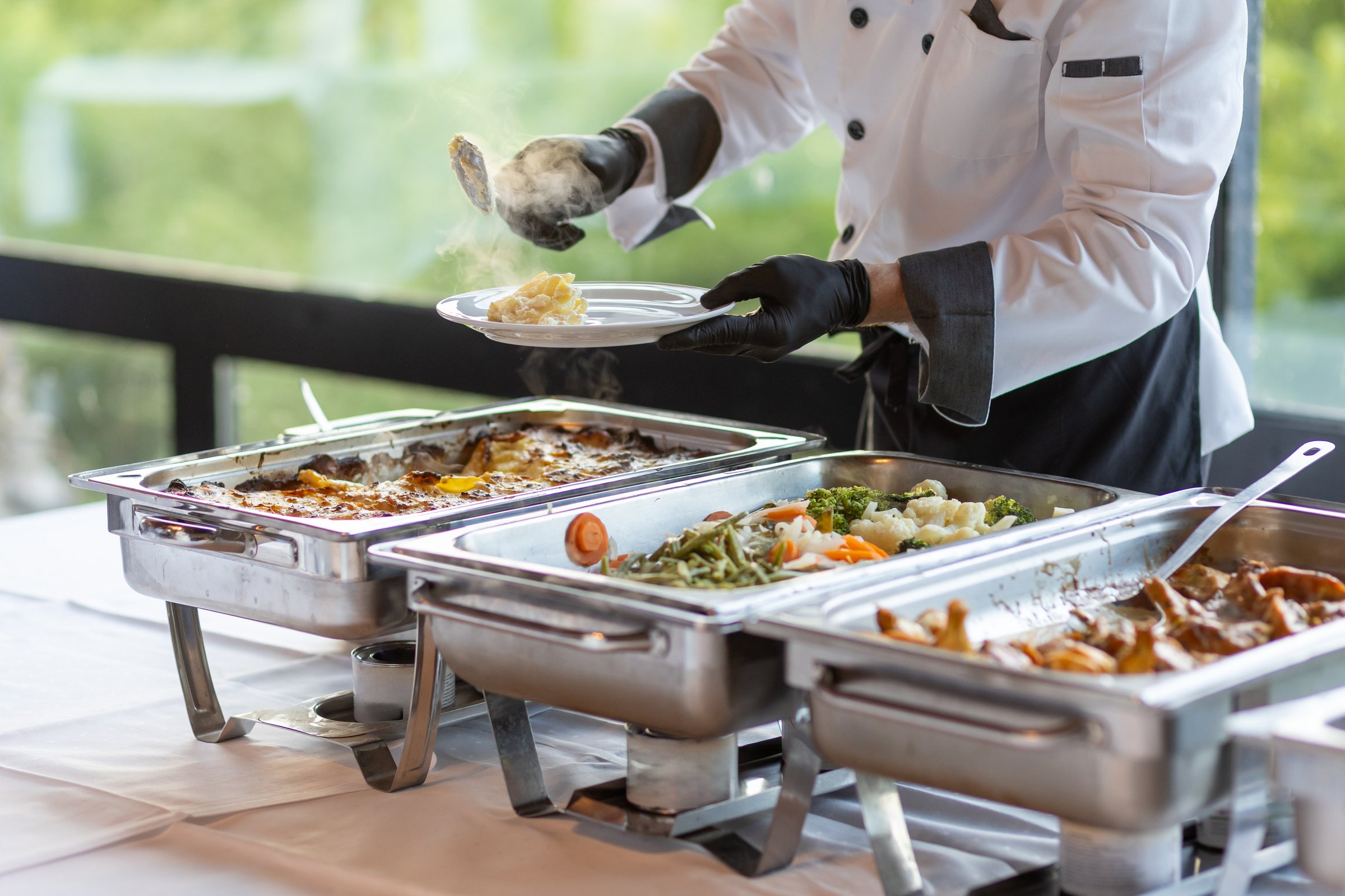 A chef in a black and white uniform expertly plates portions of food from elegant buffet trays. The setting features a bright, contemporary atmosphere with greenery in view.