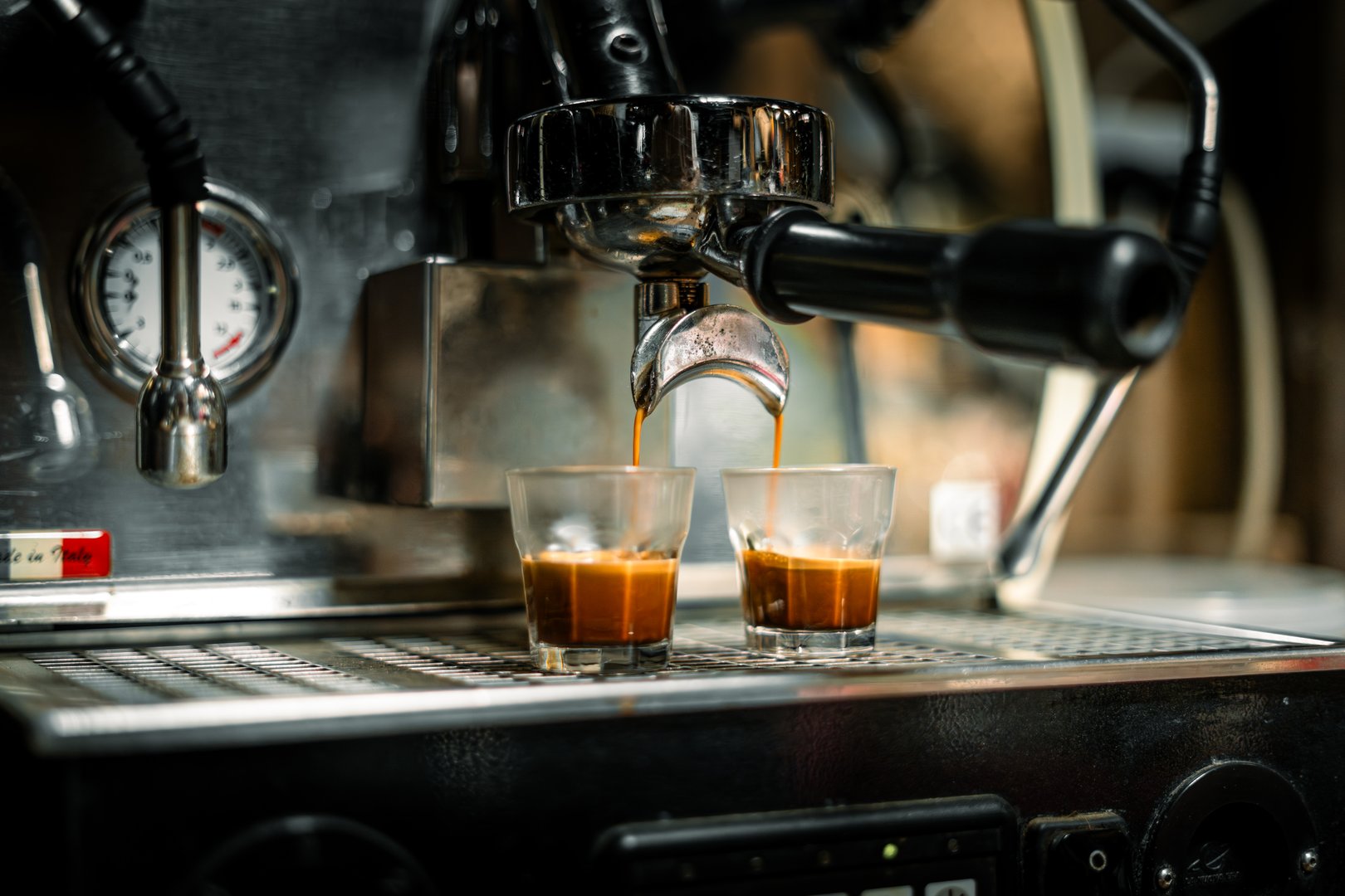 professional espresso machine in action, simultaneously brewing two shots of rich, dark espresso into small clear glasses. The golden-brown crema is visible on top of each shot