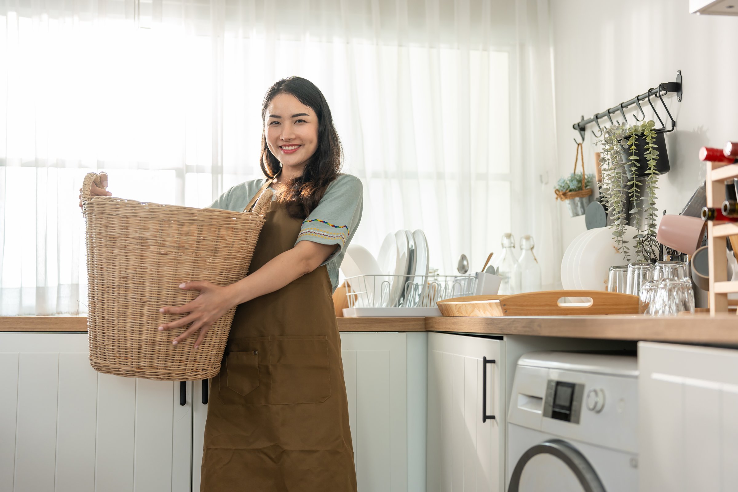 Portrait of Asian young woman doing laundry and look at camera in house. Attractive female housewife wearing apron, holding basket and prepare to put clothes to washing machine. Domestic-Housekeeping.