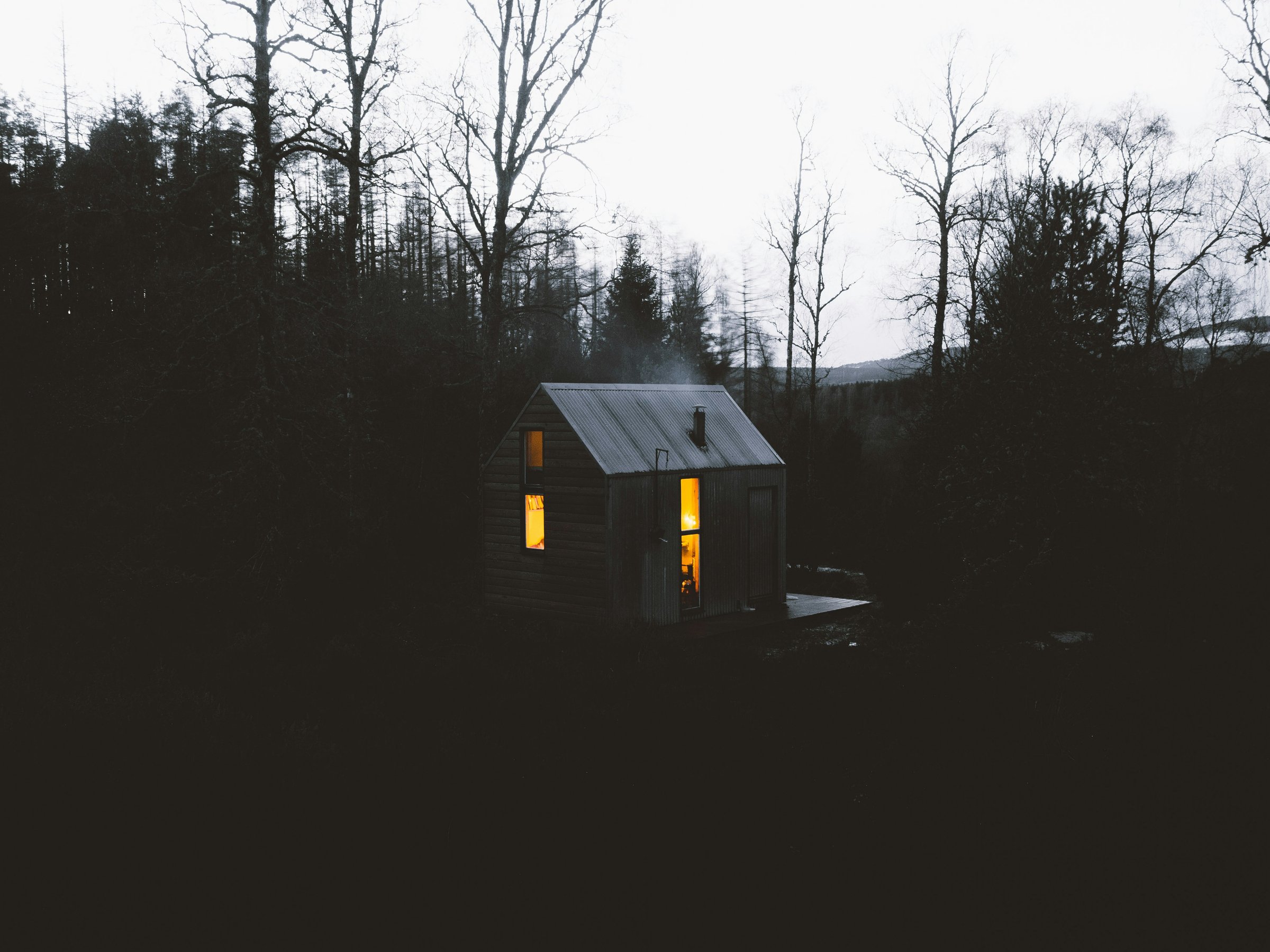 A small wooden cabin with lit windows surrounded by dark trees at dusk, creating a cozy, isolated atmosphere.
