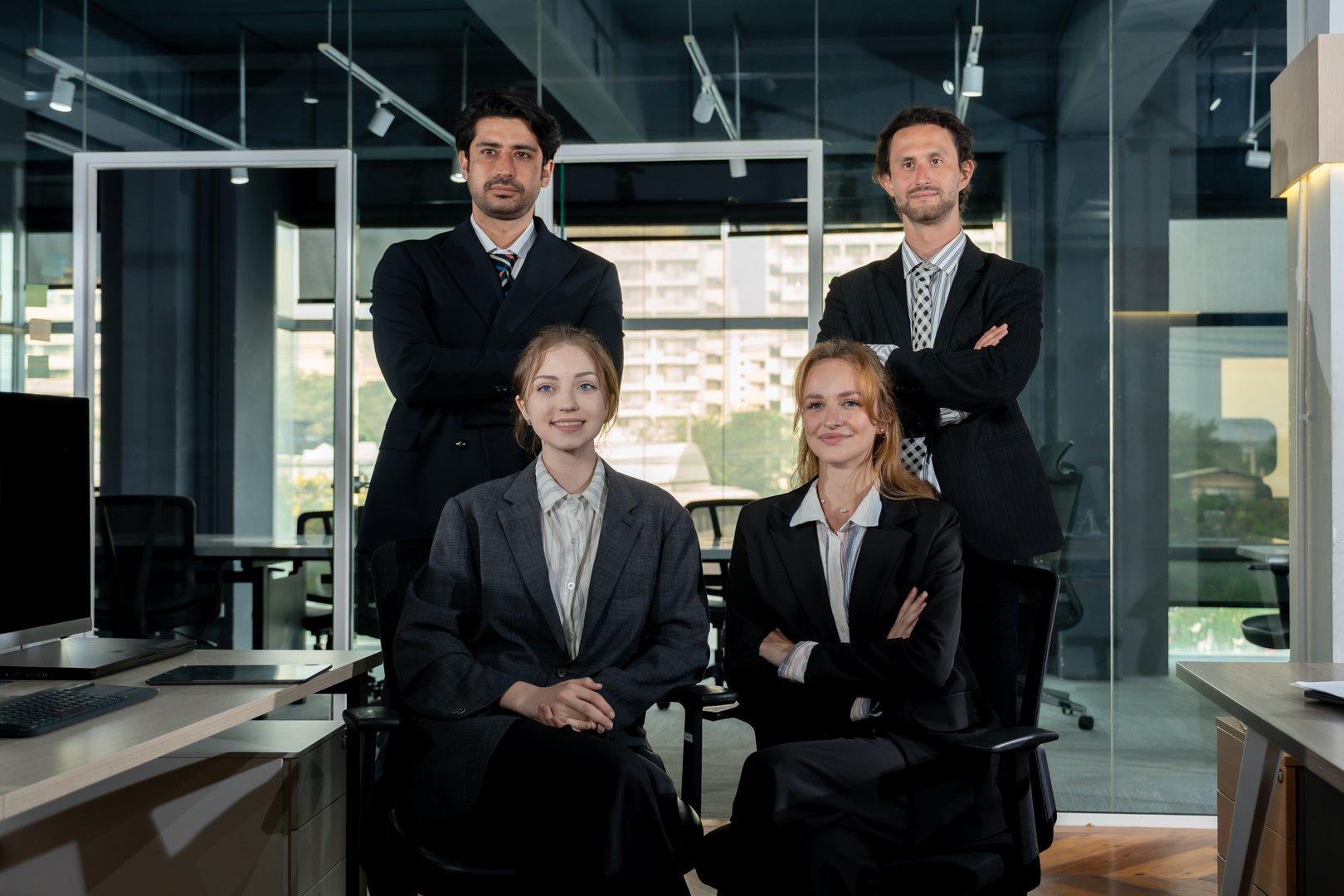 Business team posing in modern office environment. Two men standing behind two seated women, all dressed in formal attire. Confident expressions emphasize teamwork, leadership, and professionalism.