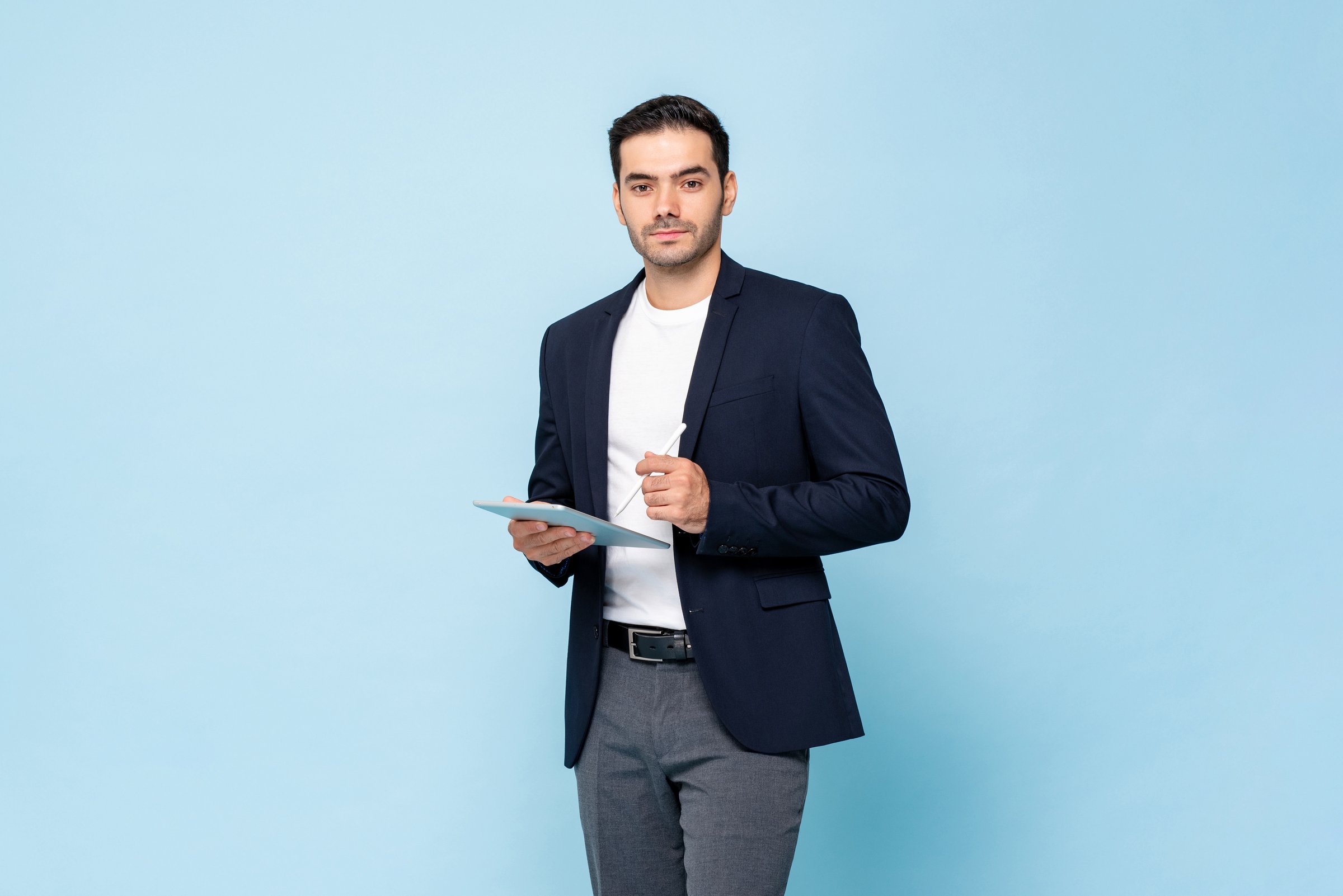 Smart handsome Caucasian man in semi formal business suit holding tablet computer with pen in light blue color  isolated studio background