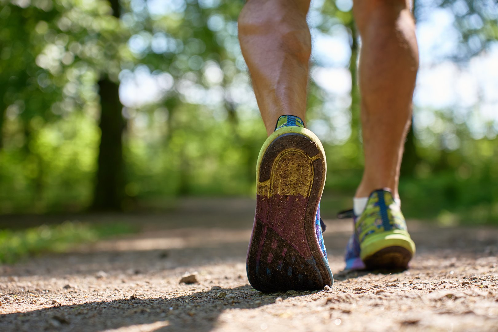 Low angle view of athletic shoes walking on sunny forest path. Close-up of male athlete running on sunny day in park. Concept of outdoor running, fitness and healthy lifestyle