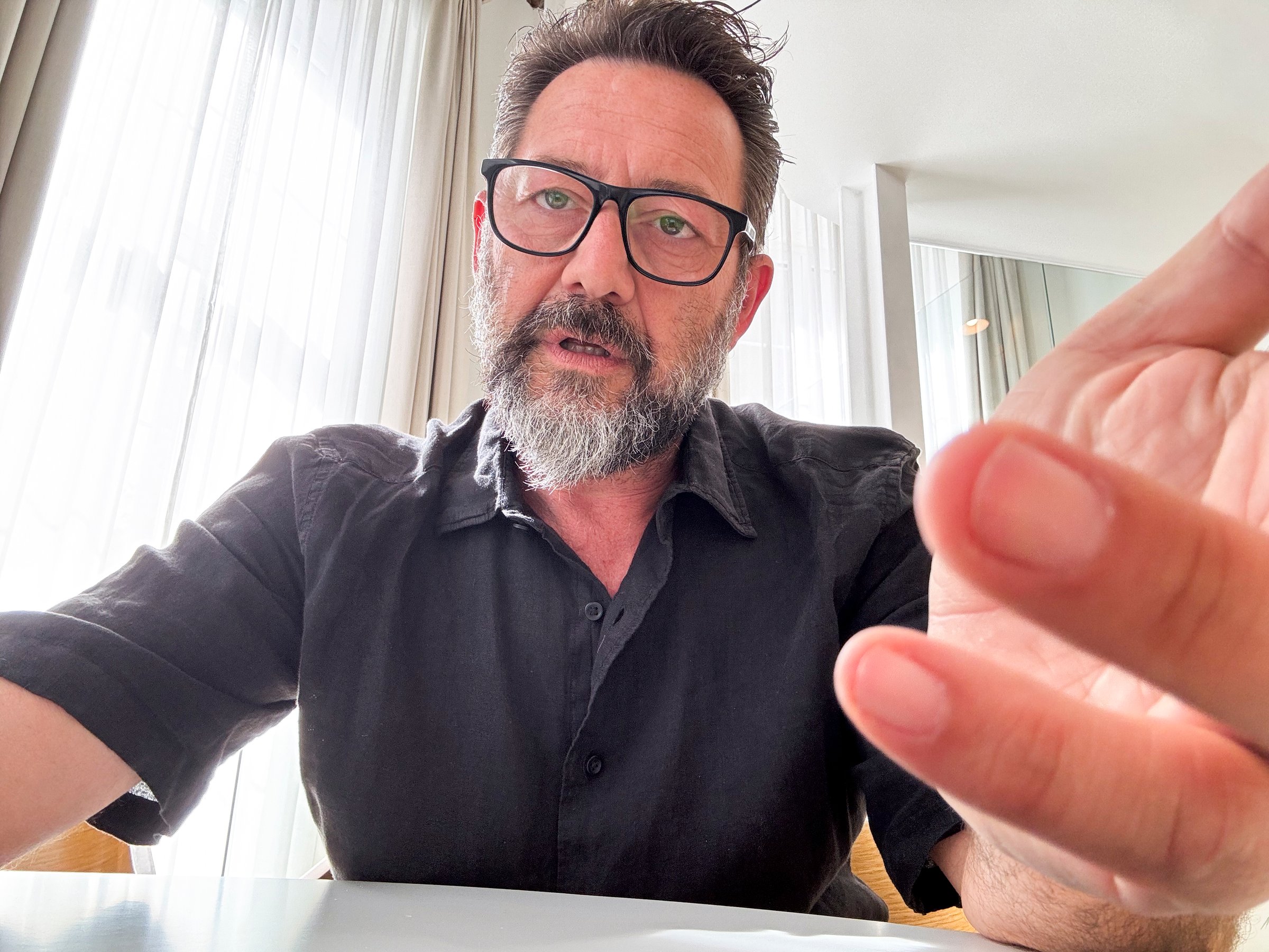 web camera view of adult man explaining online gesturing with bored expression. Businessman working in hotel room. Mature businessman wearing black glasses and talking on camera computer connection