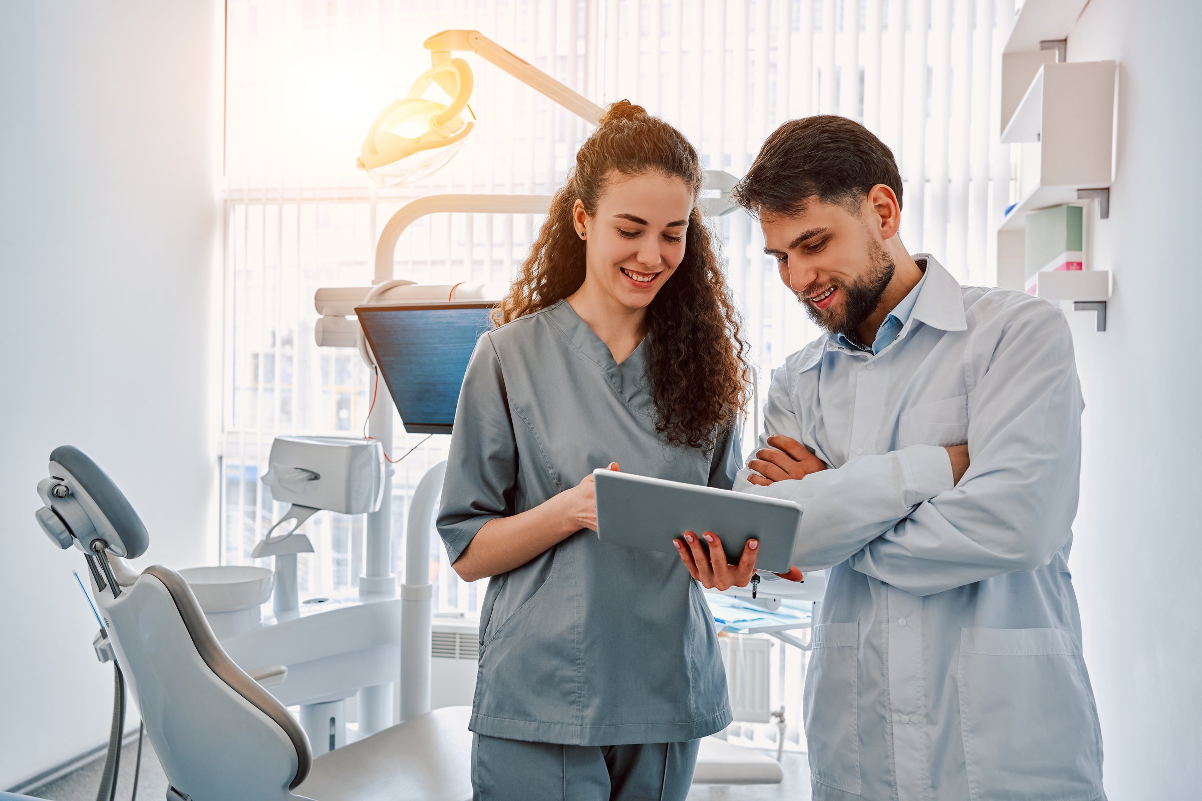 Two dentist doctors are standing in a dental office and looking at a tablet. Sunlight.