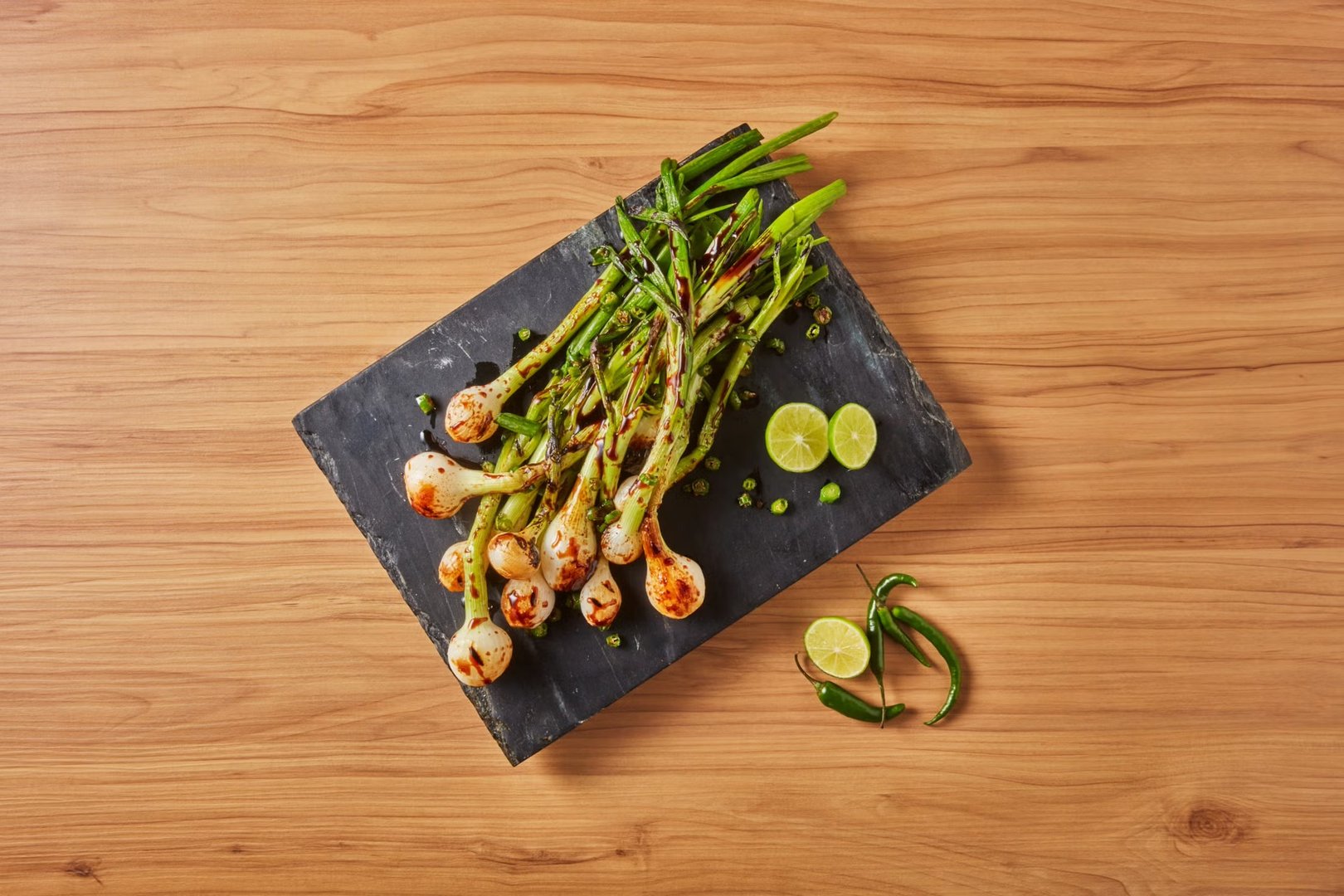 Grilled green onions on a slate board with lime slices and green chilies on a wooden surface.