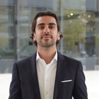 Man in a dark suit and white shirt standing in front of a glass building, looking confidently at the camera.