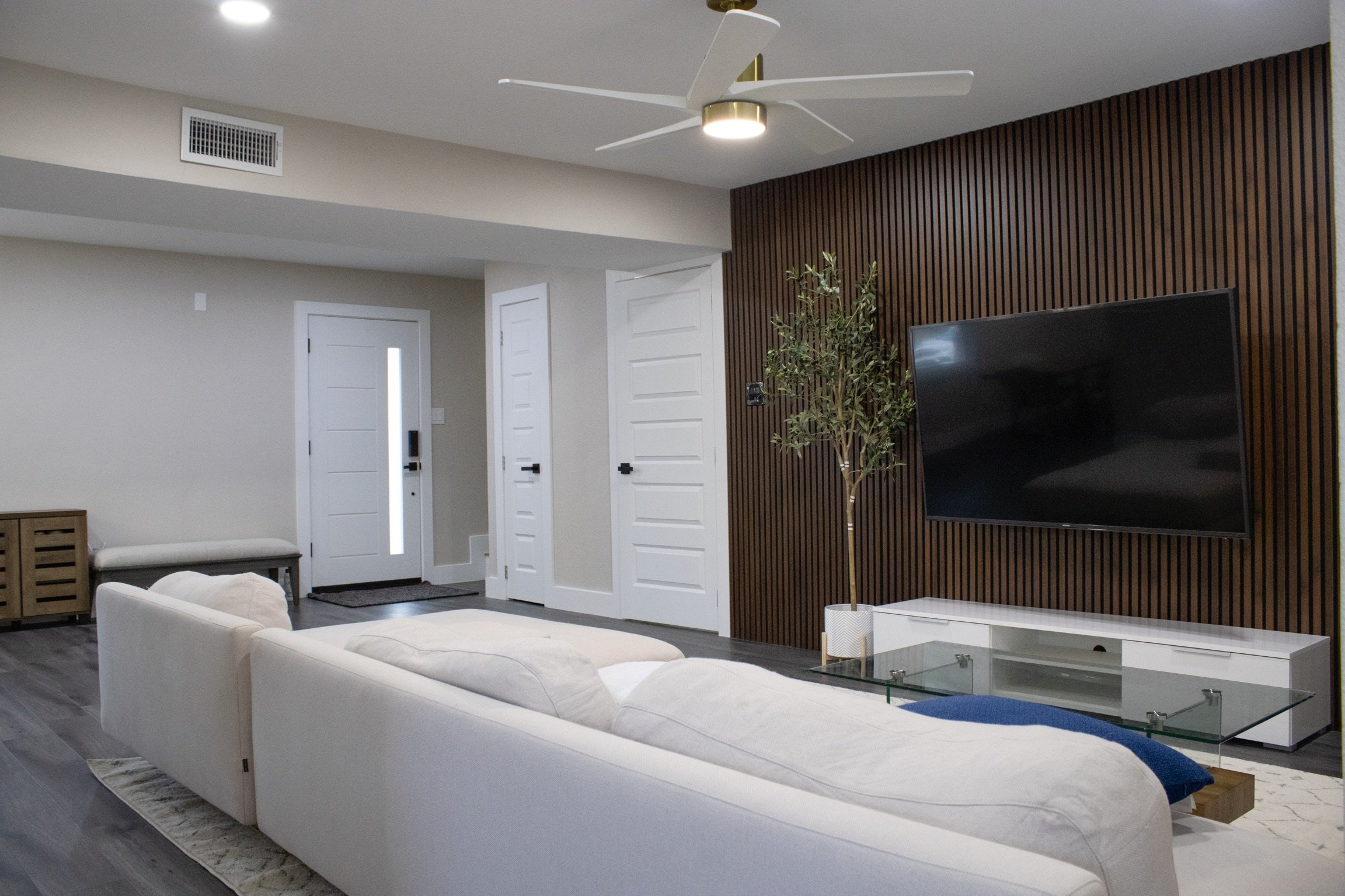 Modern living room with white sofa, large TV on a wooden slat wall, ceiling fan, and glass coffee table.