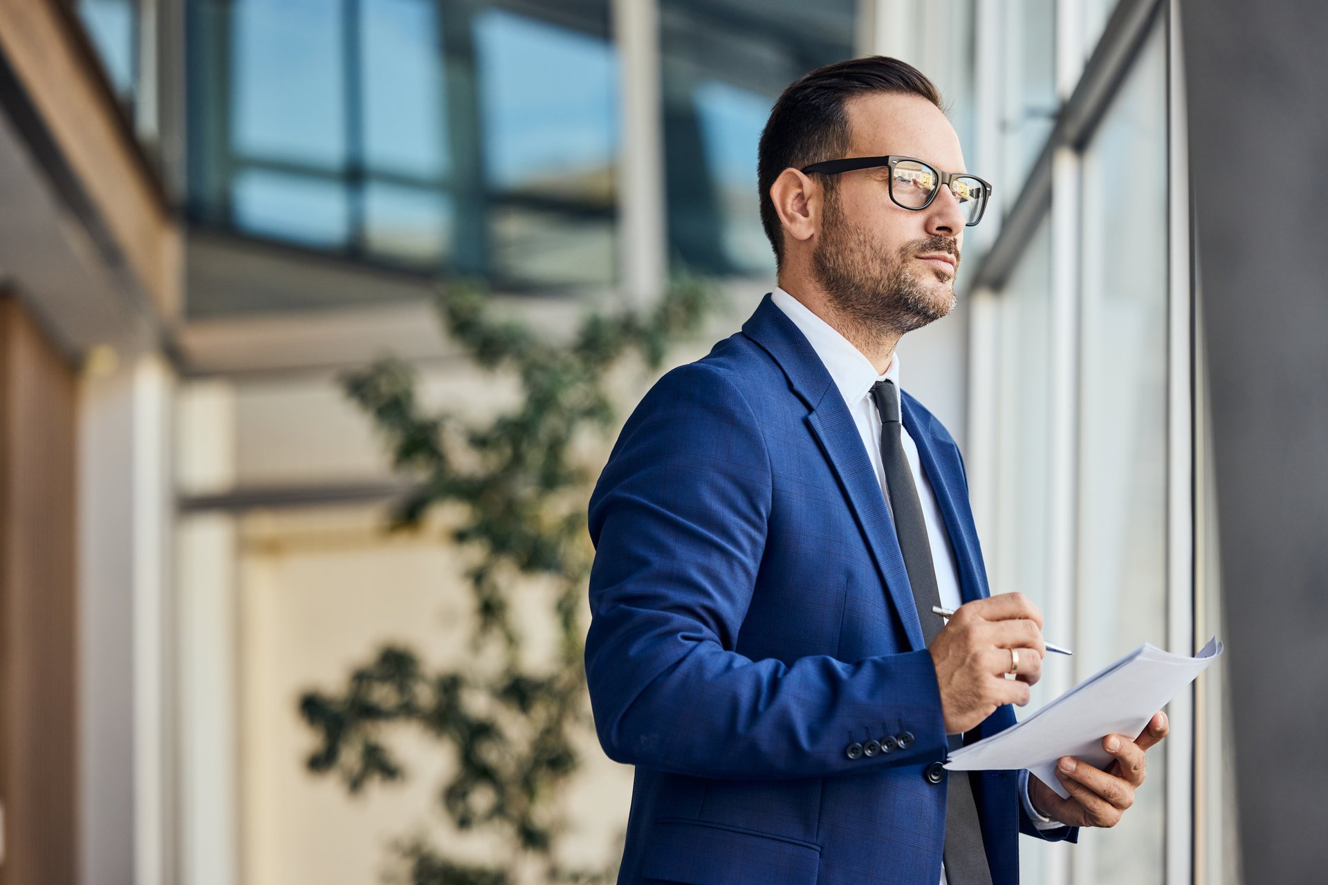 Professional businessman in modern office contemplating while holding document and pen.