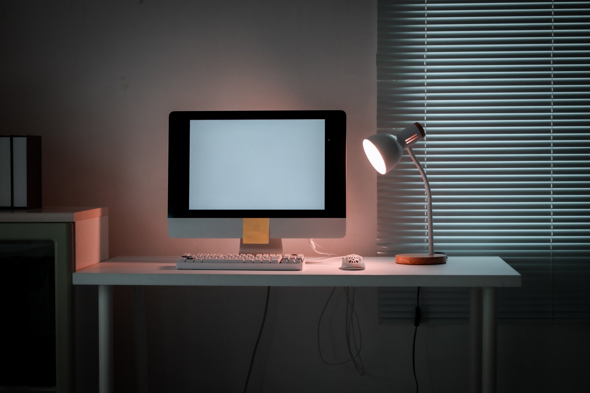 Desktop computer with a blank screen on a desk, illuminated by a glowing desk lamp. Working late or studying in a quiet, dark home office