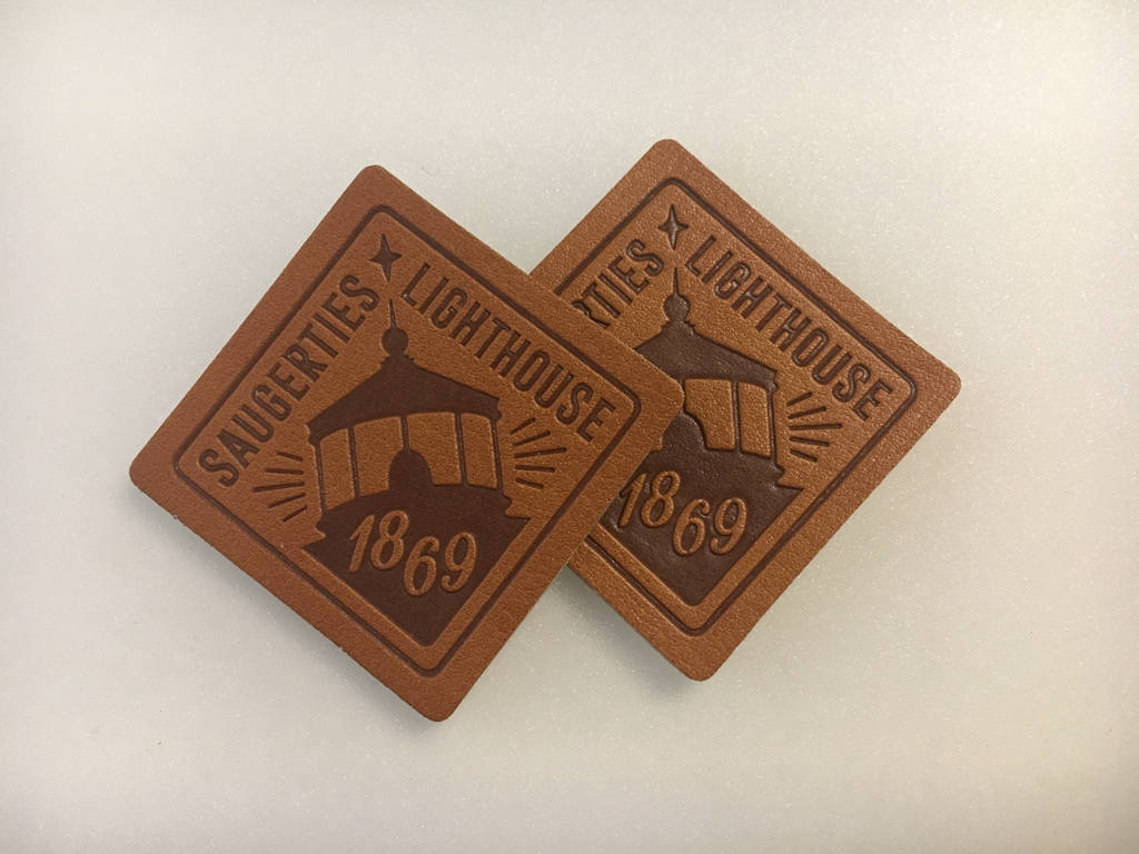 Two brown coasters with embossed Saugerties Lighthouse 1869 design featuring a lighthouse silhouette.