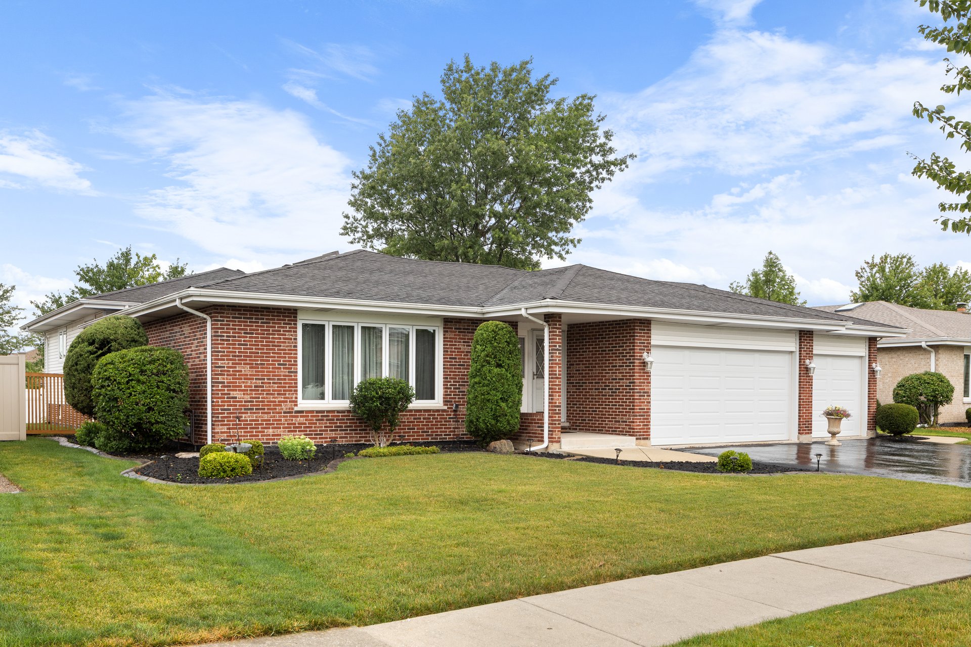 Romeoville, IL, USA - July 10, 2024: A suburban home with brick siding, a three car garage, neat landscaping, and a wet blacktop driveway.