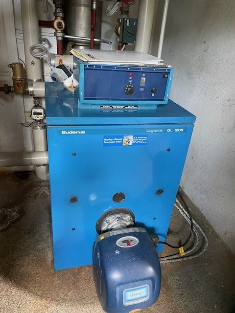 A blue Buderus Logano G205 boiler unit with an attached control panel in a utility room with exposed pipes.