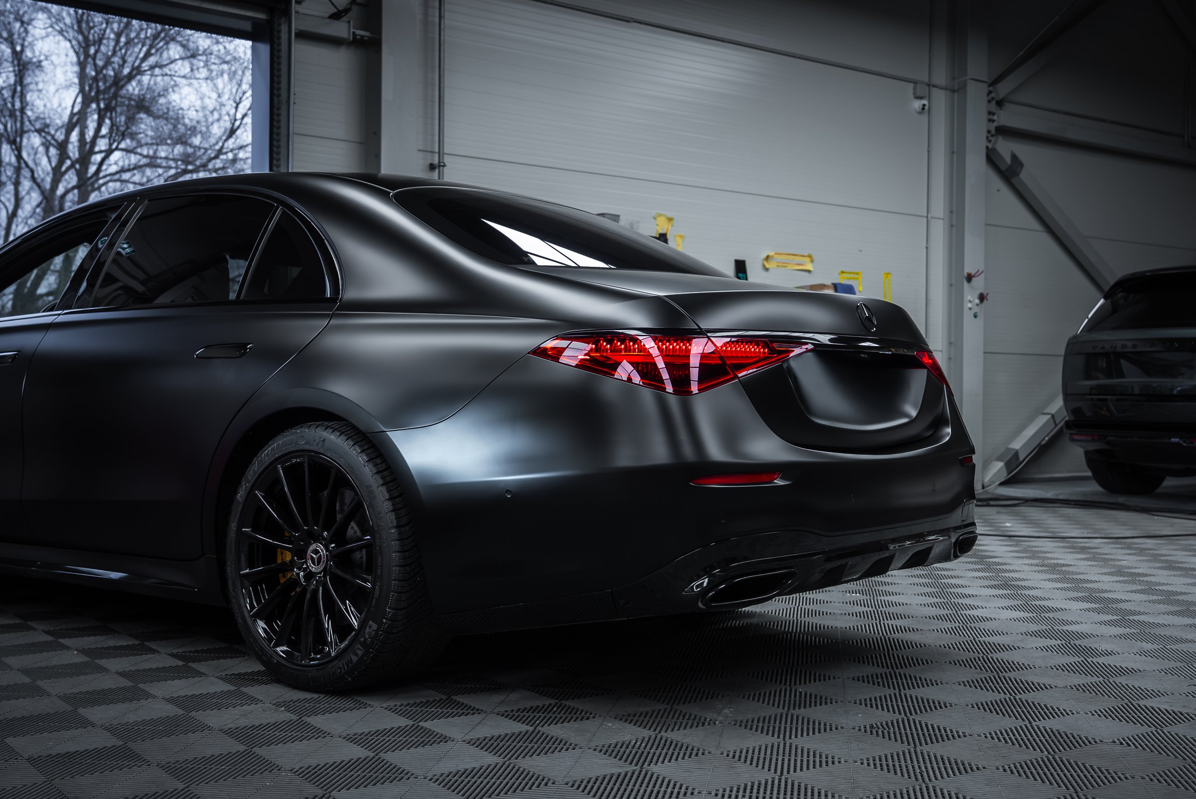 Rear three quarter view shows OLED tail lights, quad exhaust tips, and black multi spoke wheels in a detailing garage with taped fixtures and a dark SUV in back.
