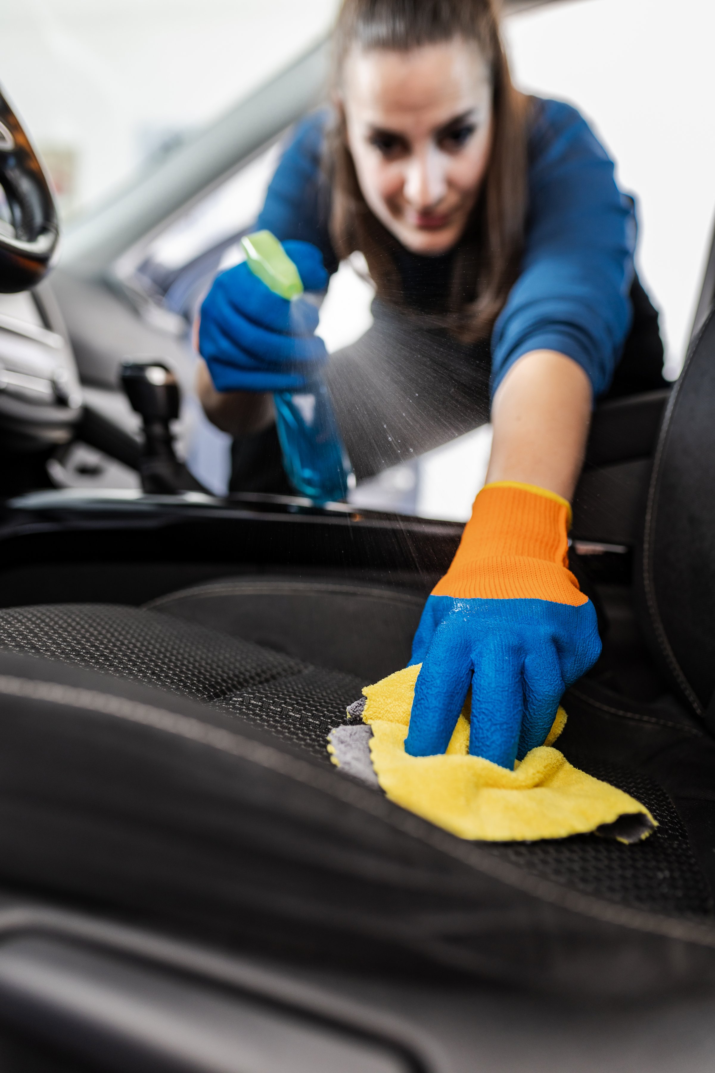 In a well-lit car, an individual in blue shirt and orange gloves is diligently cleaning the seats with a spray cleaner and a cloth, ensuring every corner sparkles.