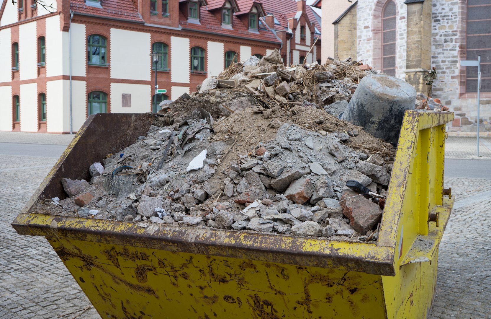 A yellow metal container filled with construction debris stands in a square in front of historic buildings