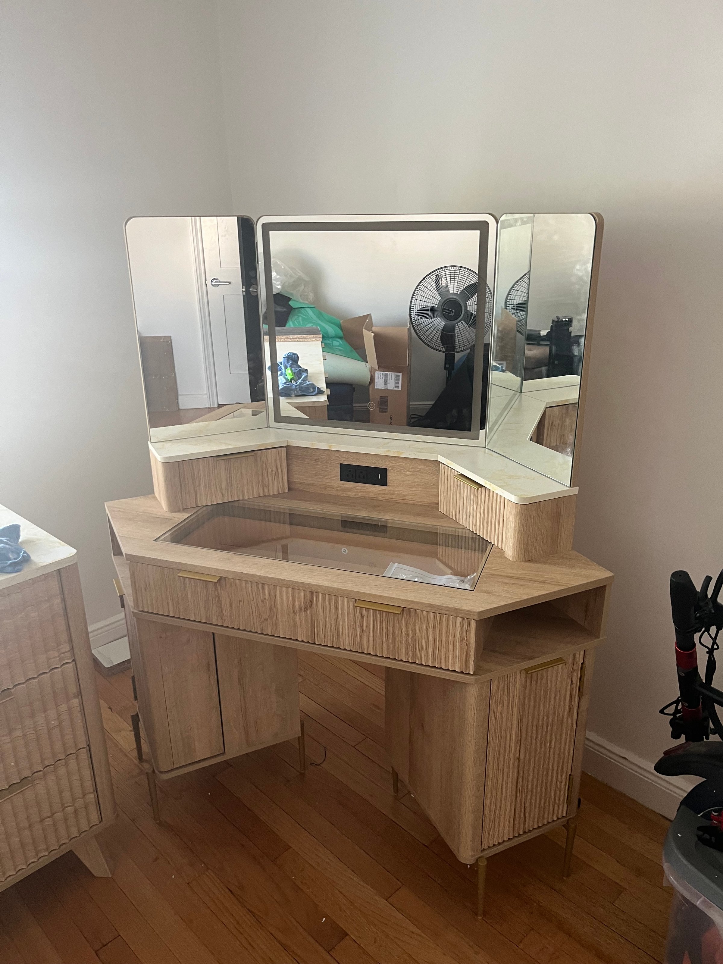 A wooden vanity with three mirrors, drawers, and brass handles stands on a hardwood floor in a room with miscellaneous items.