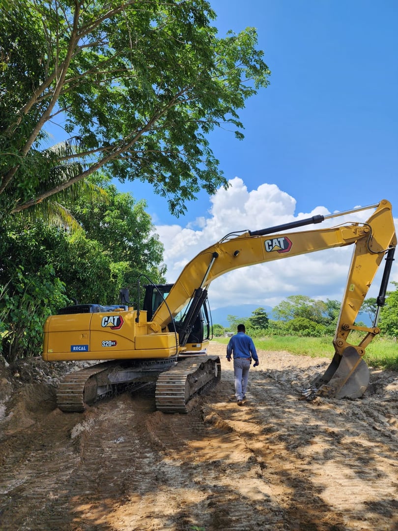 A yellow excavator parked on a dirt road under trees, with a person walking nearby, on a sunny day.