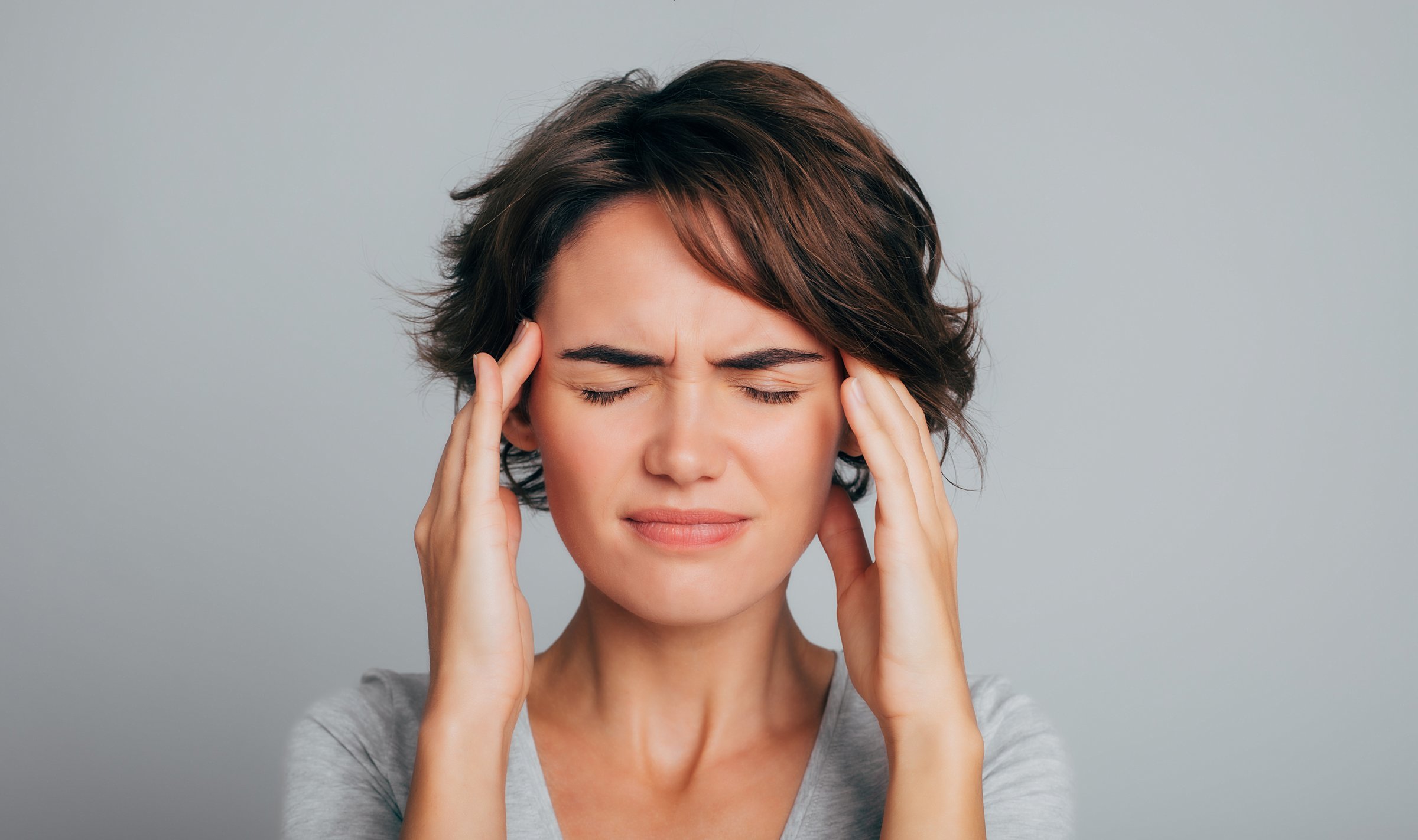 Close up photo of young woman feeling headache, migraine or being ill with short hair in t-shirt touching her head with pain isolated over gray background. Depressed and overworked