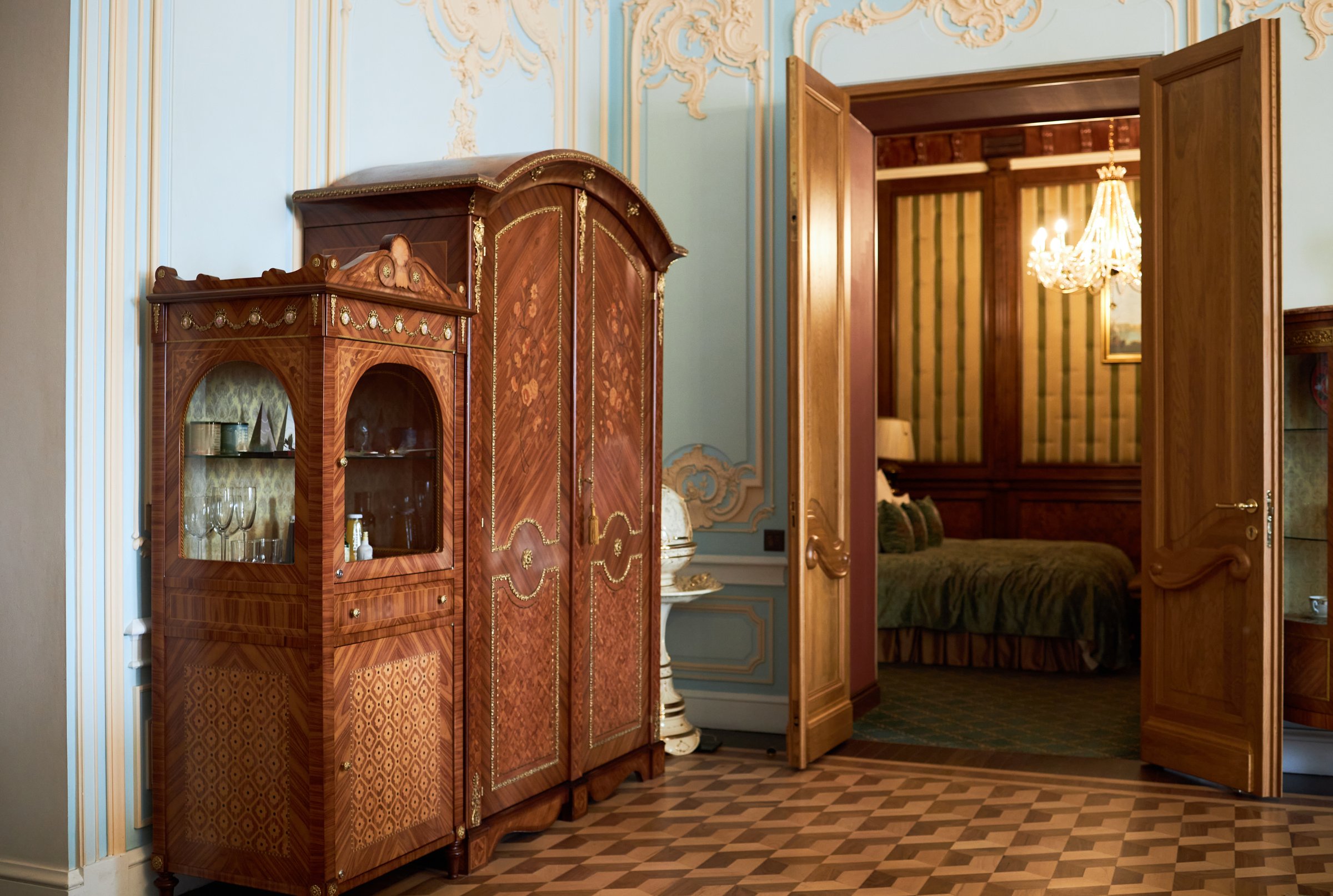Vintage wooden cabinet and armoire standing in ornate room with open door revealing glimpse of elegant bedroom and chandelier, classic parquet flooring visible in foreground
