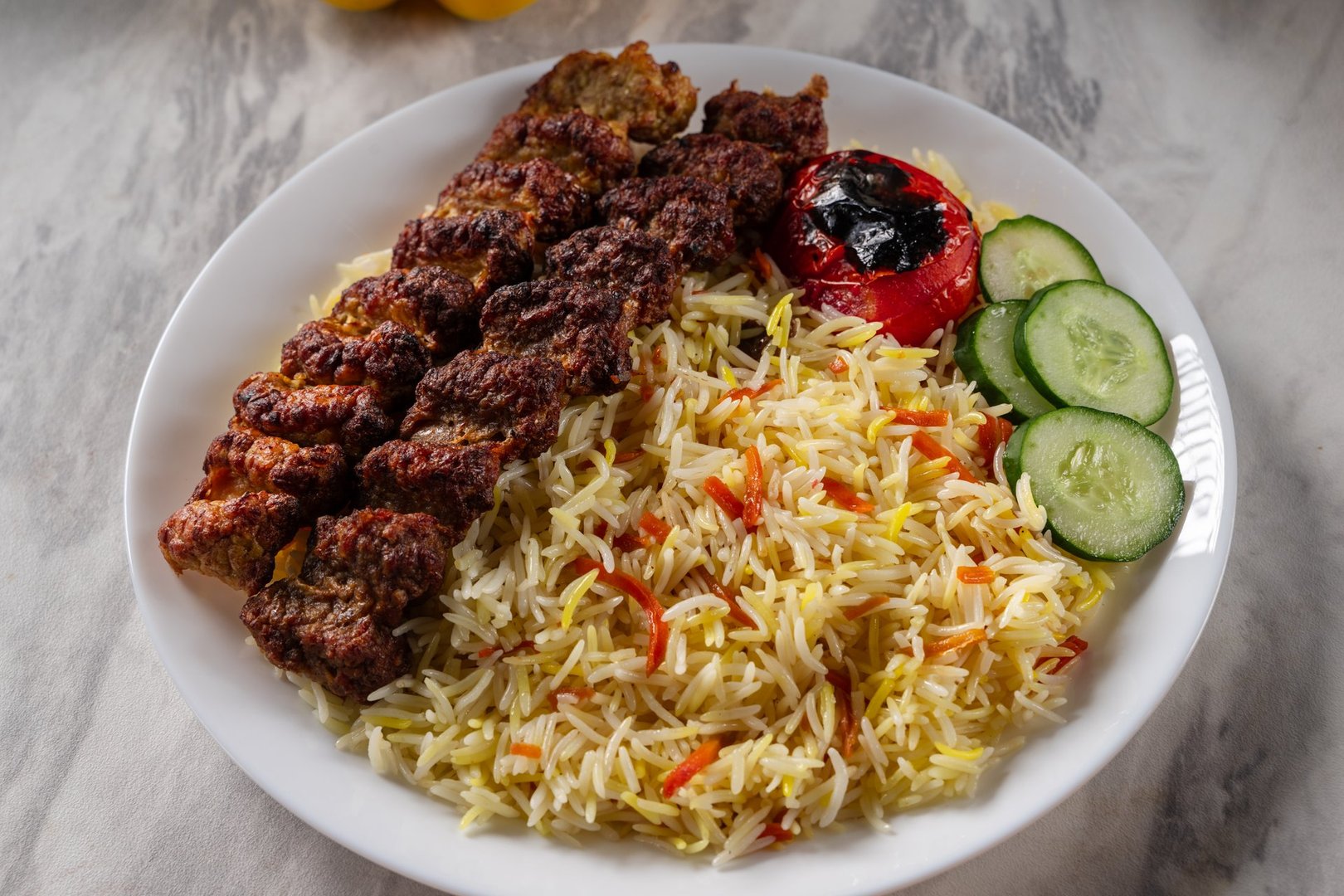 Grilled beef kebab served with colorful basmati rice, slices of fresh cucumber, and a roasted tomato on a white ceramic plate, placed on a marble surface in natural light