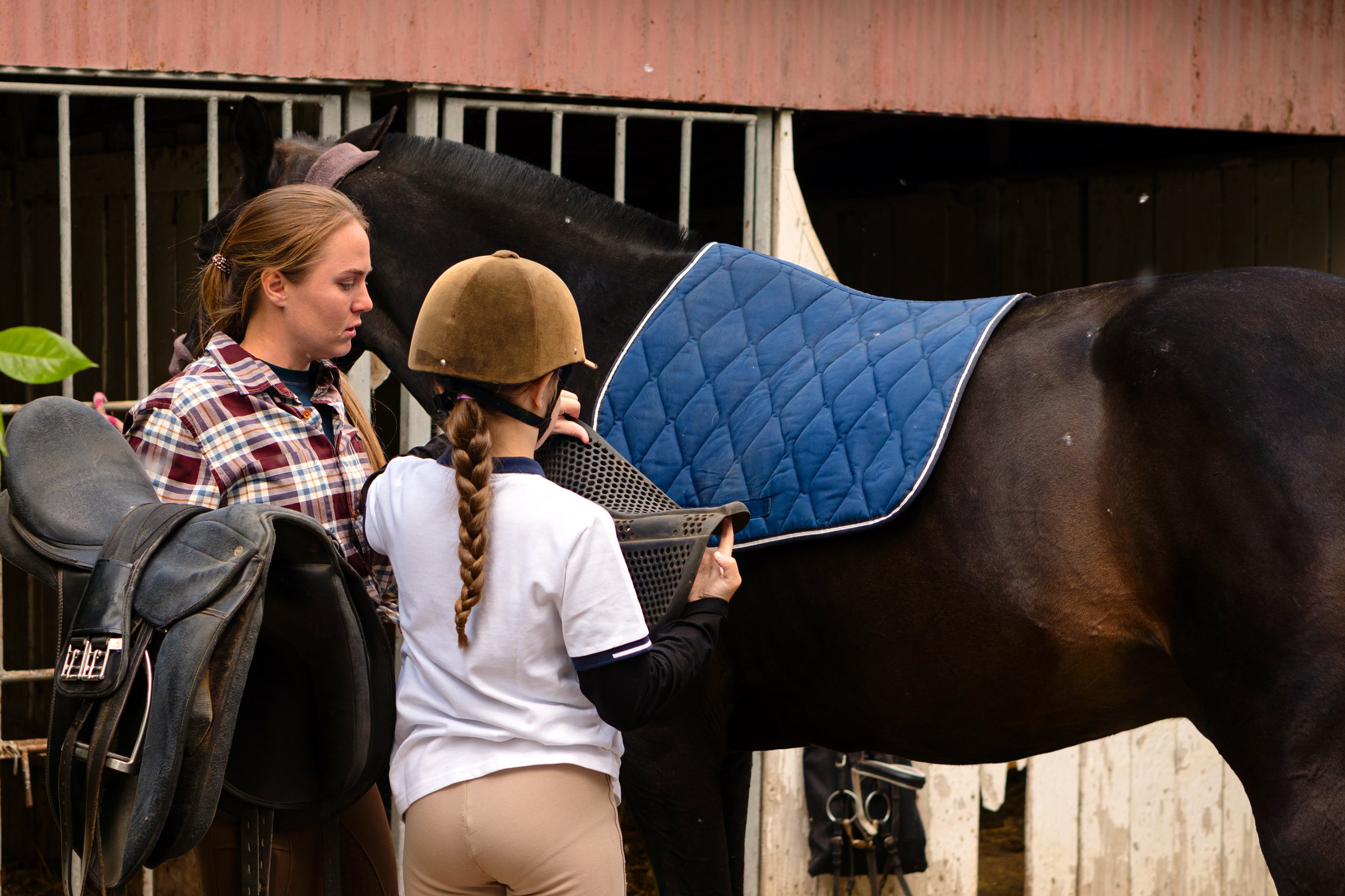 Instructor helping a young rider girl in safety helmet and polo shirt position a saddle on a horse at a stable.
