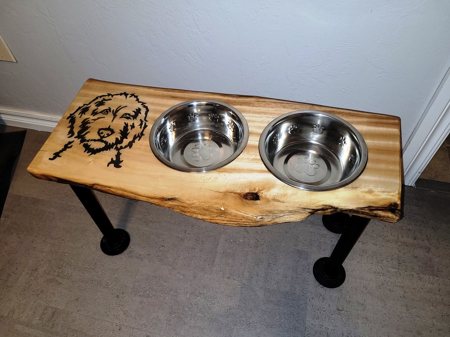Wooden dog feeding station with metal legs and two stainless steel bowls, featuring a dog face engraving on the surface.