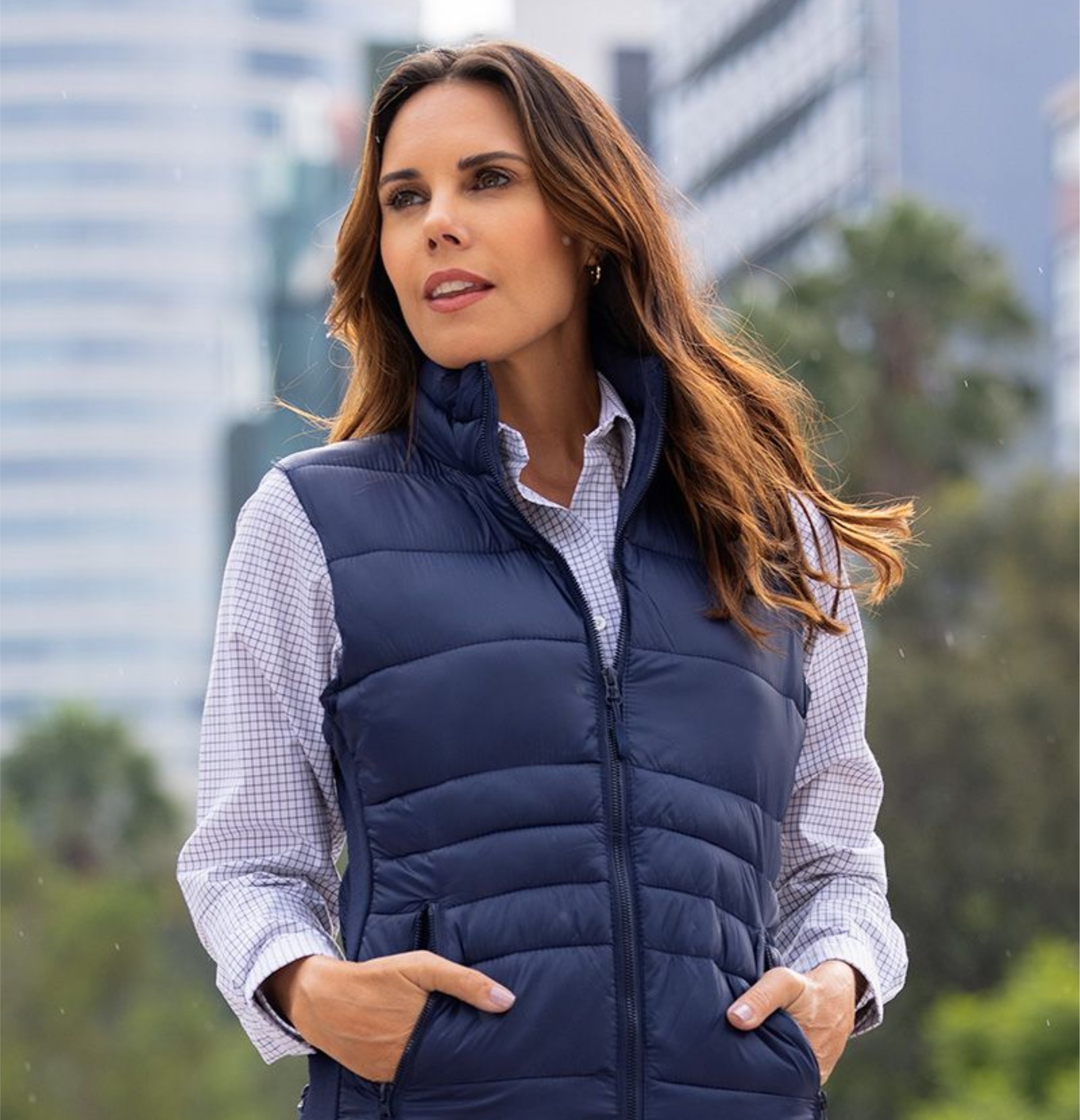 Woman in a navy puffer vest and checkered shirt stands outdoors, with city buildings and trees in the background.