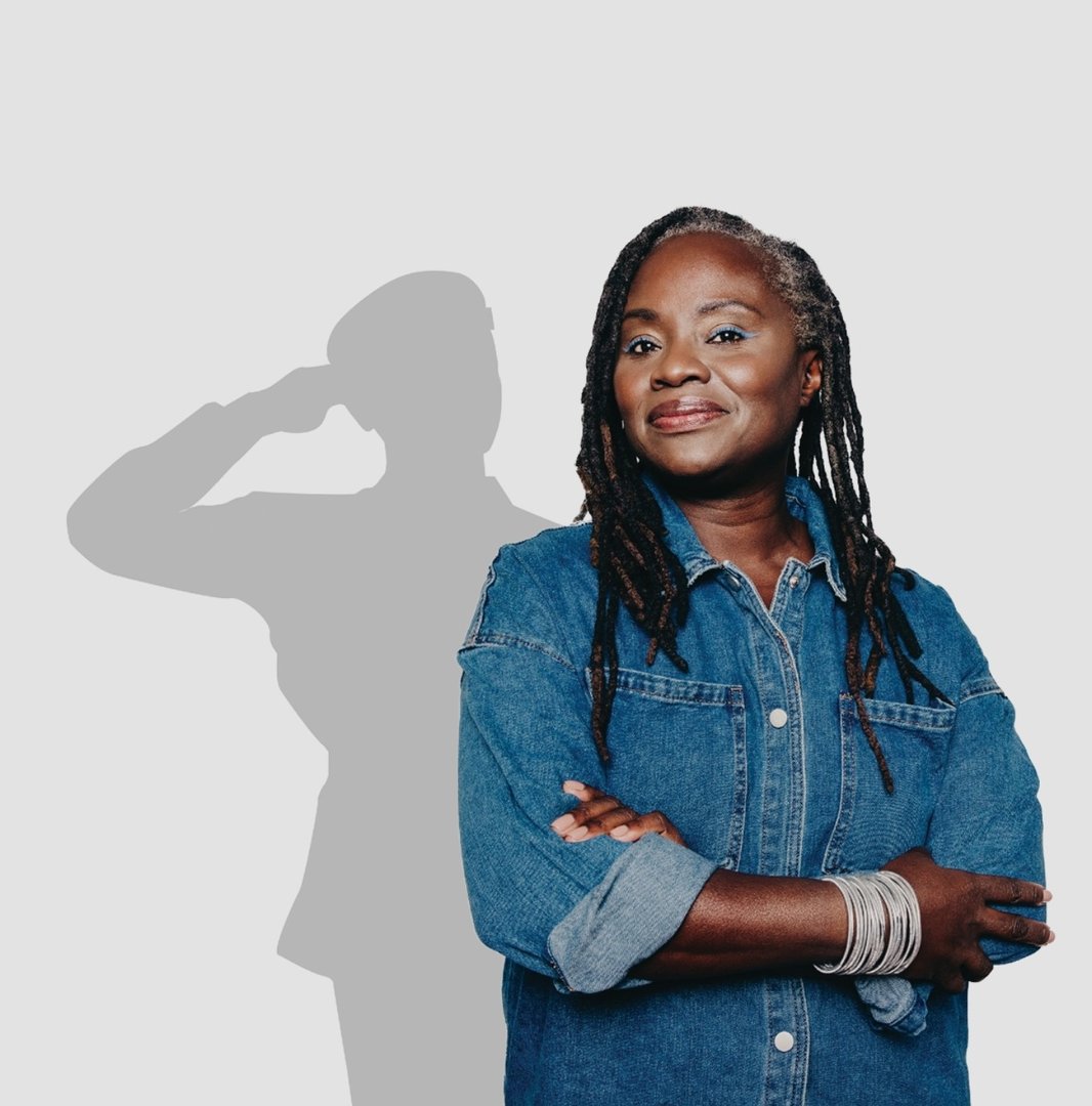 Woman in a denim jacket smiles with arms crossed, standing in front of a silhouetted soldier saluting.