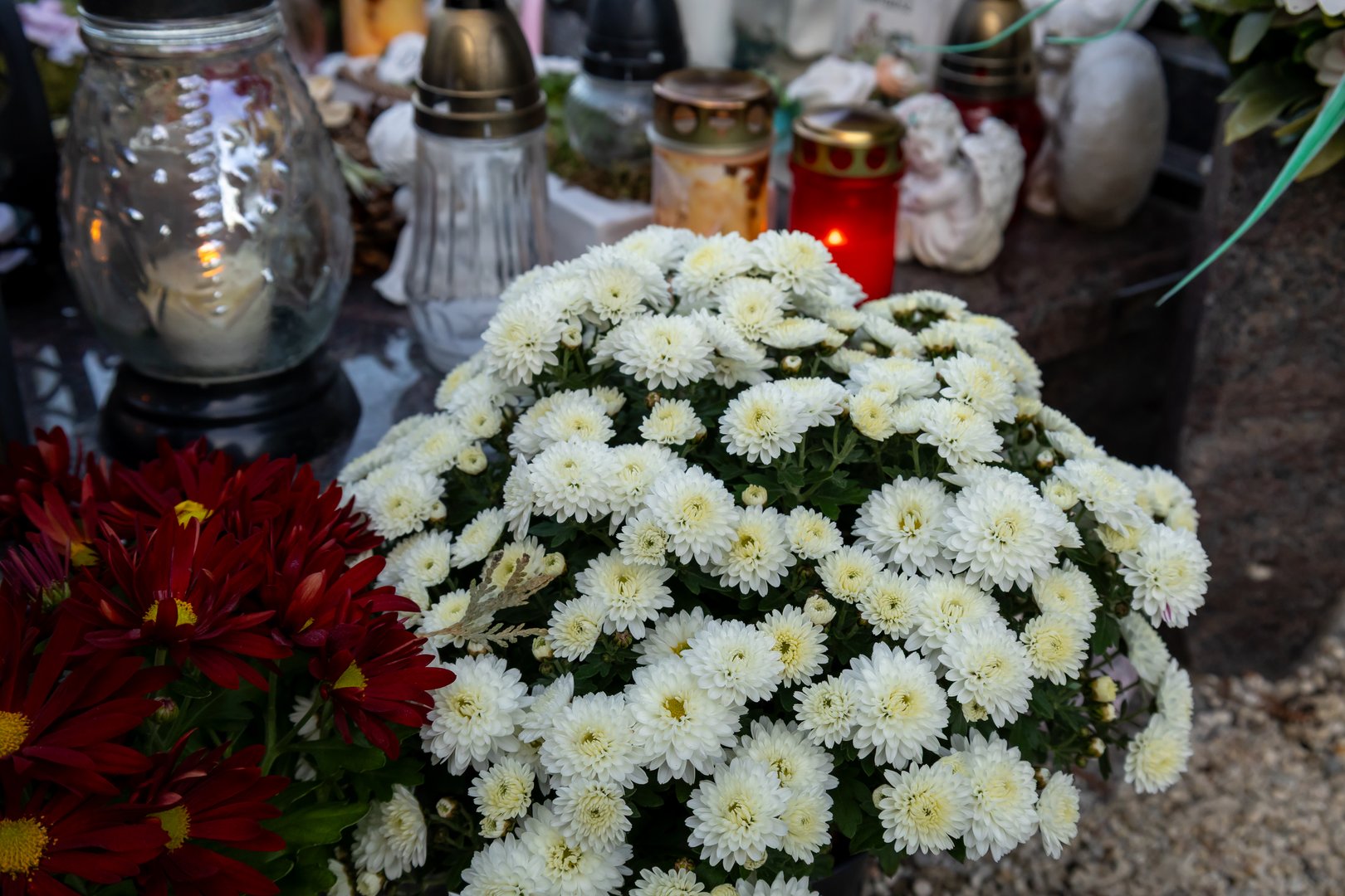 White chrysanthemum flowers arranged near memorial lanterns and burning candles in a cemetery setting. The simple floral tribute and quiet surroundings reflect mourning and heartfelt remembrance for departed loved ones.