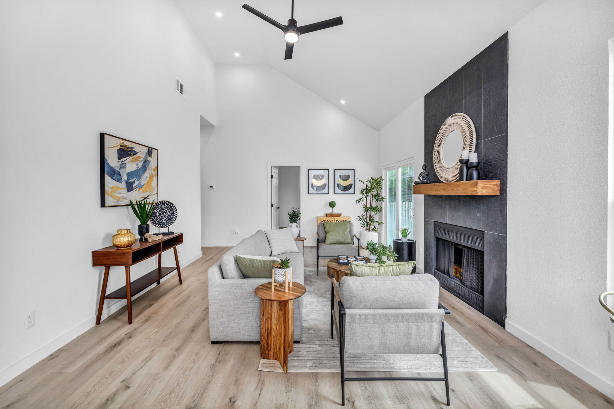 Modern living room with a gray sofa set, wooden accents, abstract artwork, and a black tile fireplace with a round mirror.