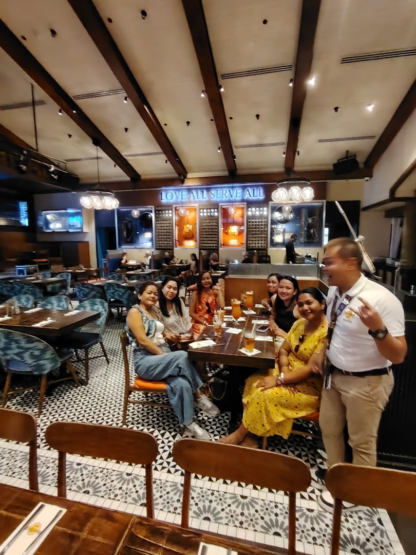 A group of people sitting around a table in a restaurant with drinks, under a sign that reads "Love All, Serve All."