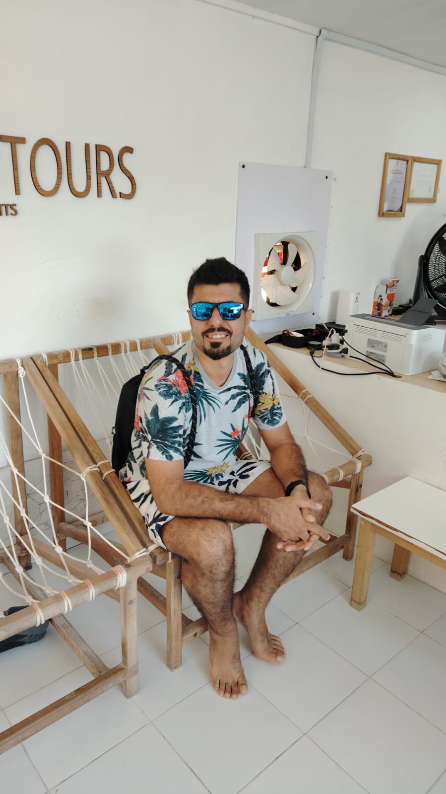 Man in sunglasses and floral shirt sitting on a wooden hammock seat indoors next to an office setup.