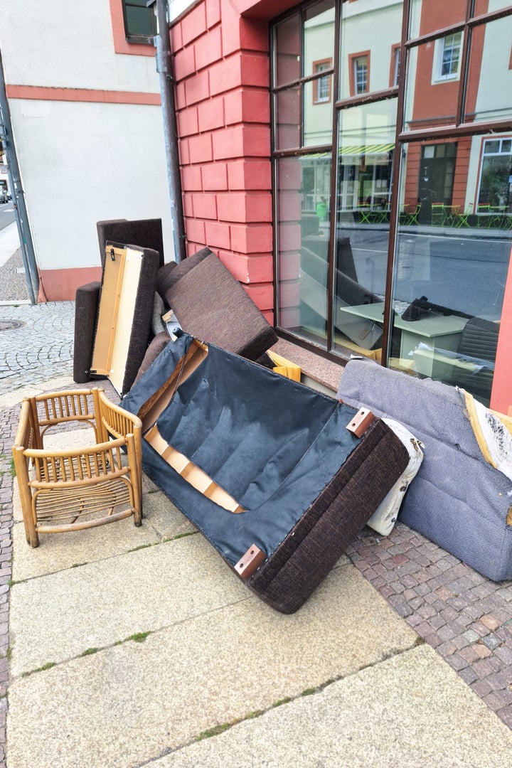 A pile of discarded furniture including a sofa, chair, and various cushions on a city sidewalk, highlighting urban waste and the need for proper disposal or recycling. Vertical picture