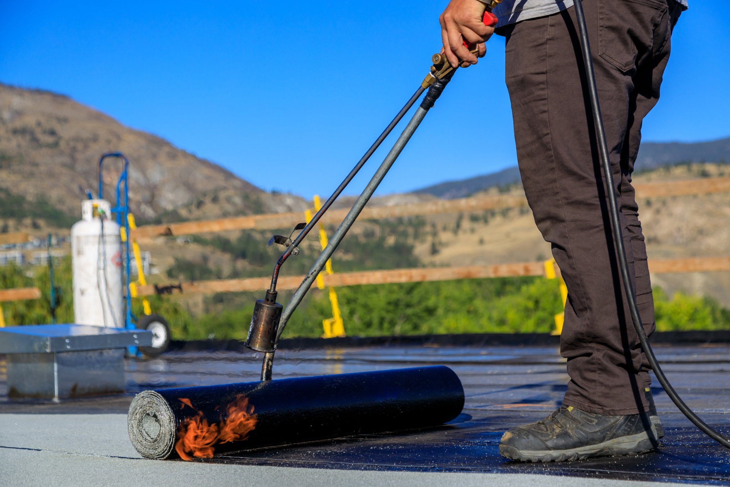 A roofer torches a membrane layer on a flat roof using a tiger torch to seal the asphalt bitumen, ensuring a durable waterproof layer.