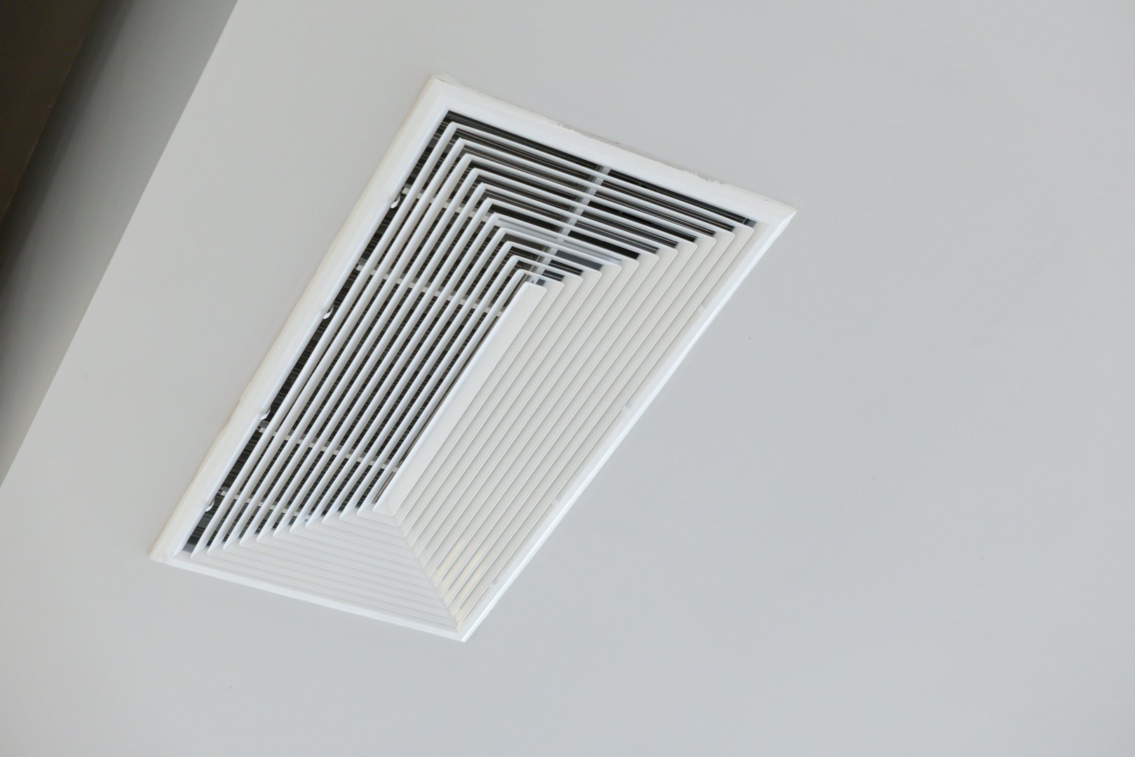 A white vent with black stripes is on a white wall. The vent is angled up and is the only thing visible in the image