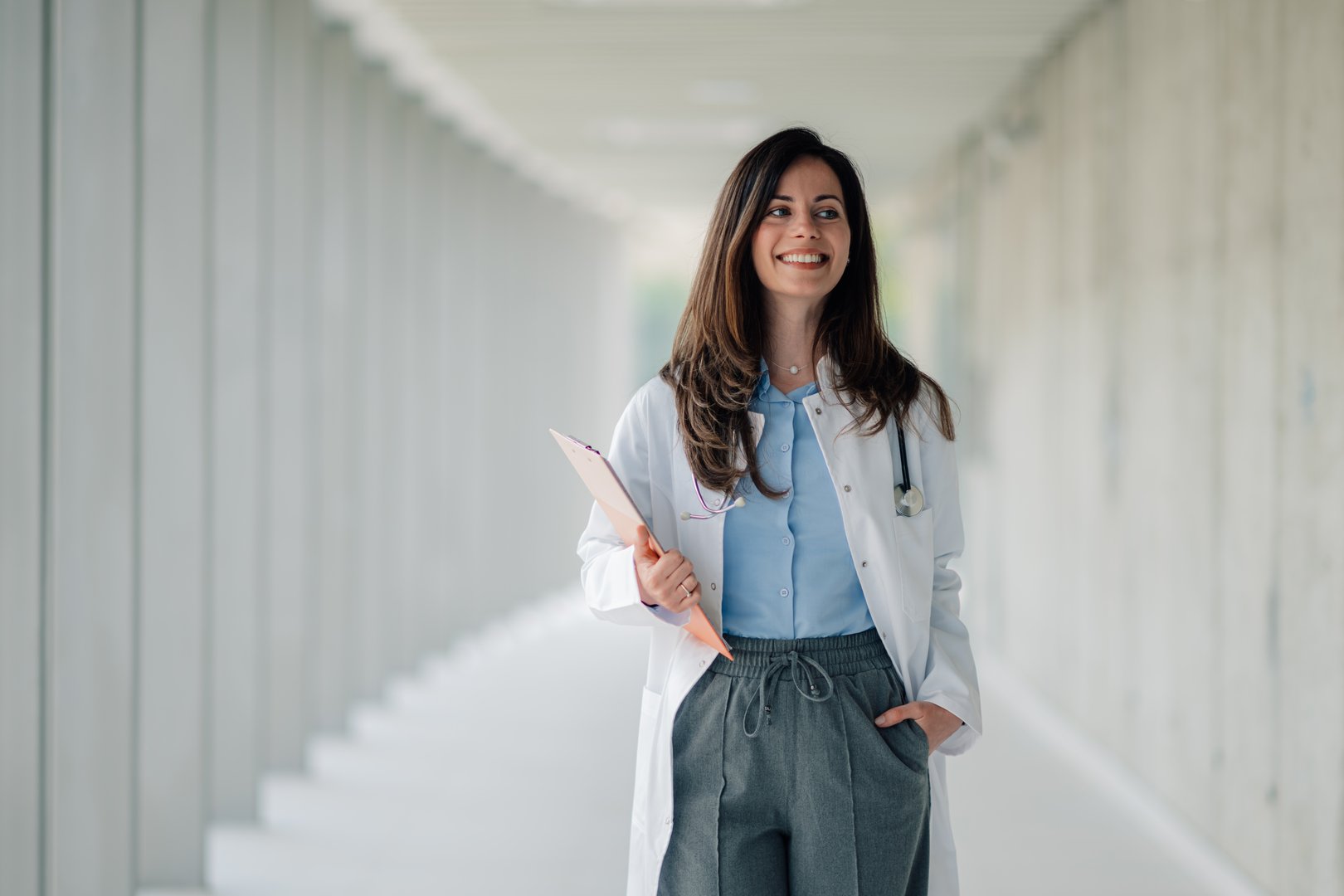 Young female doctor smiling and walking through a modern hospital corridor holding a clipboard
