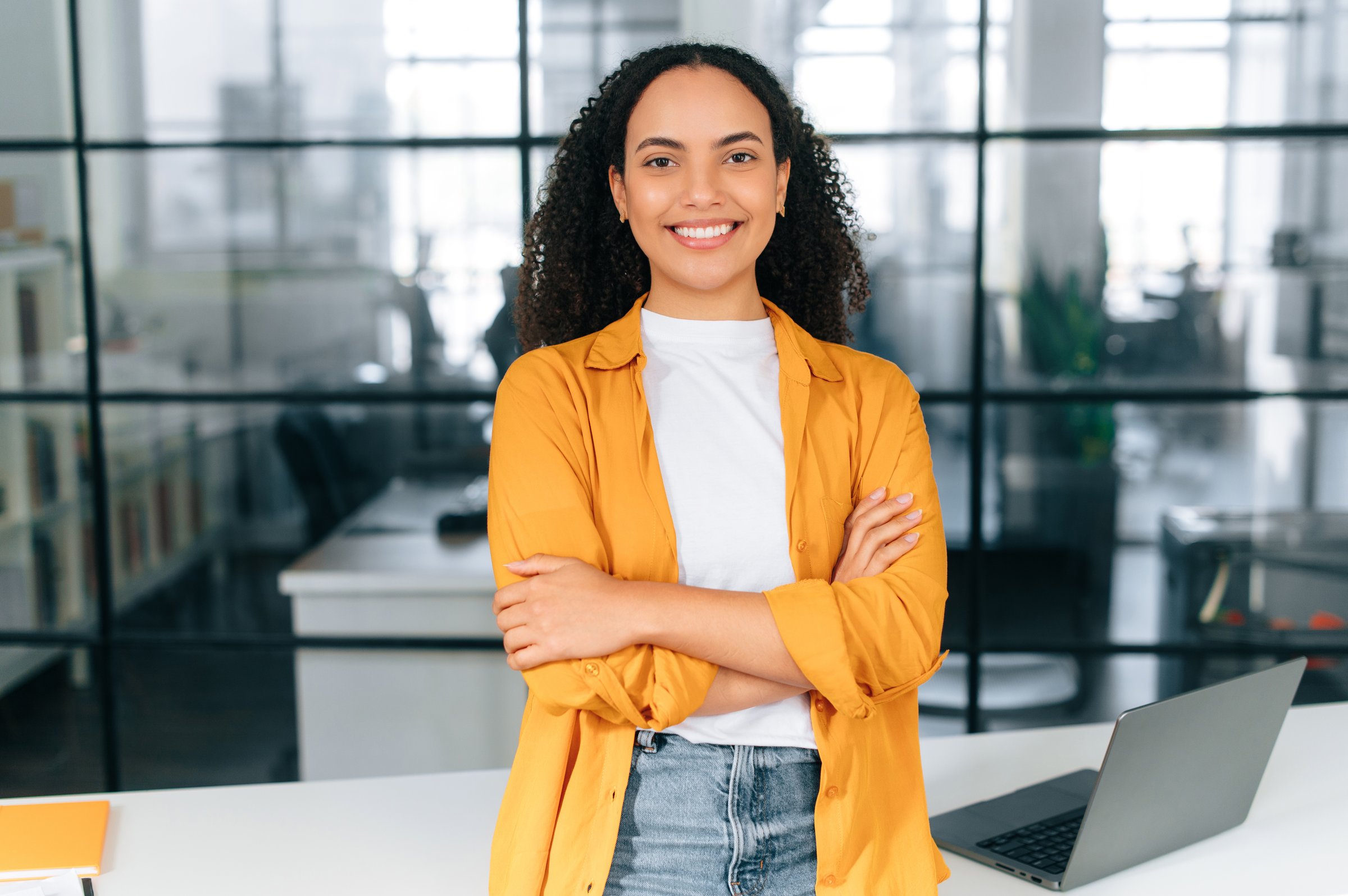 Proud successful gorgeous hispanic or brazilian curly haired young woman in a casual stylish clothes, stand in the modern office near work desk with arms crossed, looks at camera, smiling happily