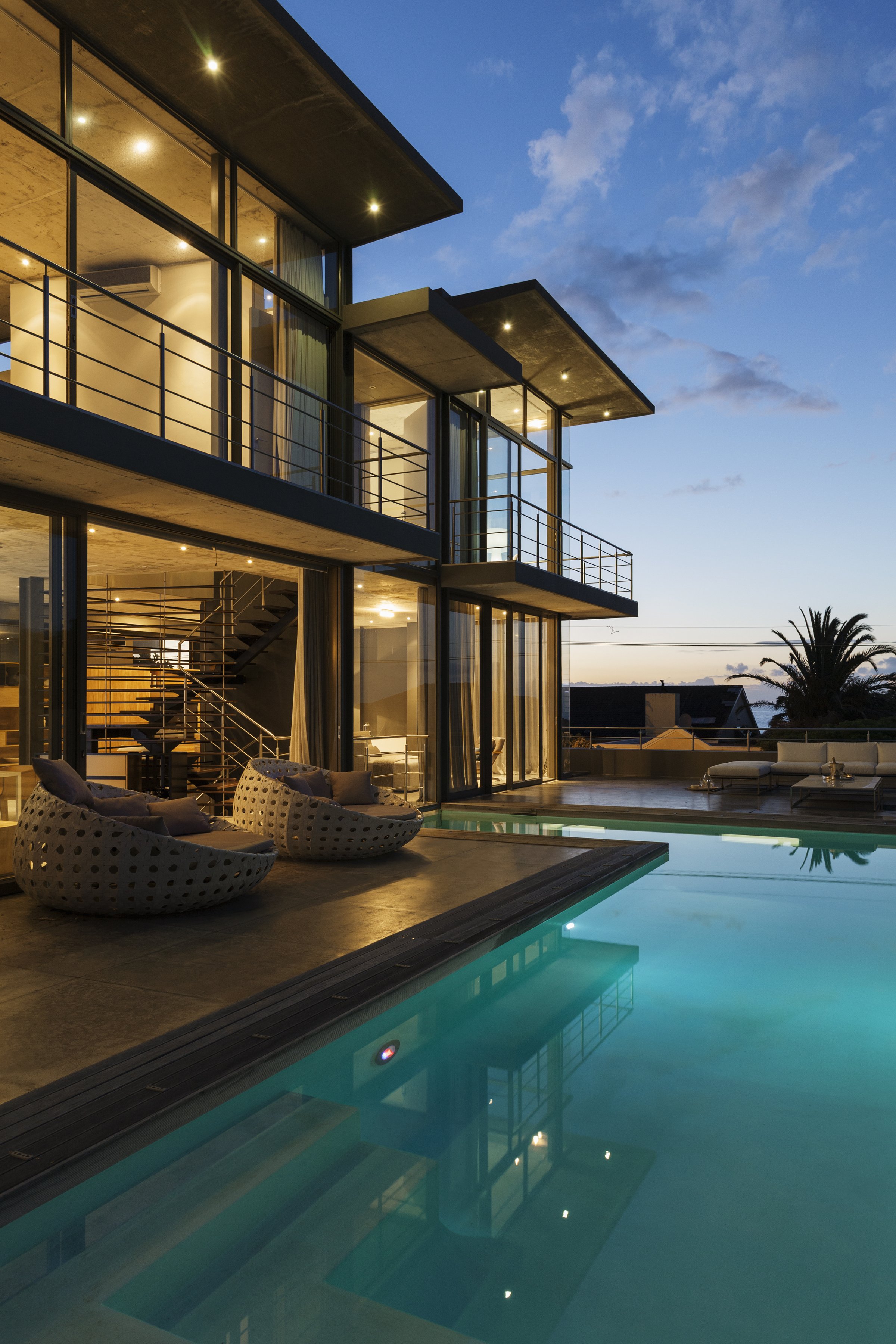 Modern house with large windows, poolside seating, and illuminated pool at dusk, showcasing sleek architectural design.