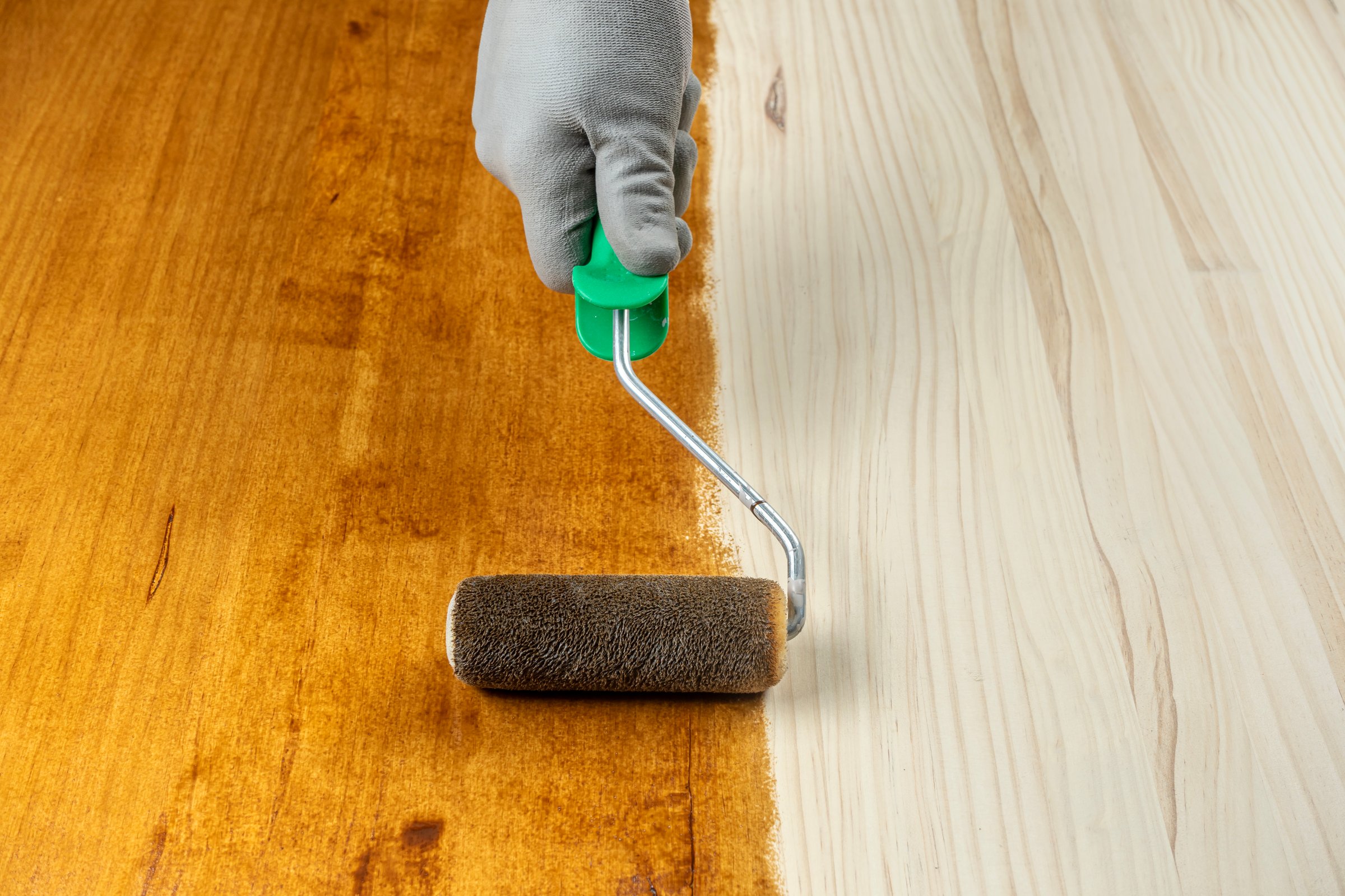 Hand wearing glove applies wood stain with roller on wooden surface, showing contrast between stained and natural wood.