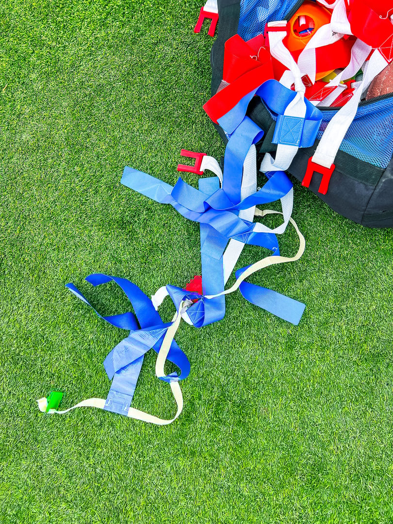A large athletic duffel bag is filled with equipment for flag football play. Red, blue and green flags and a football overflow from the bag. Green football field turf background.