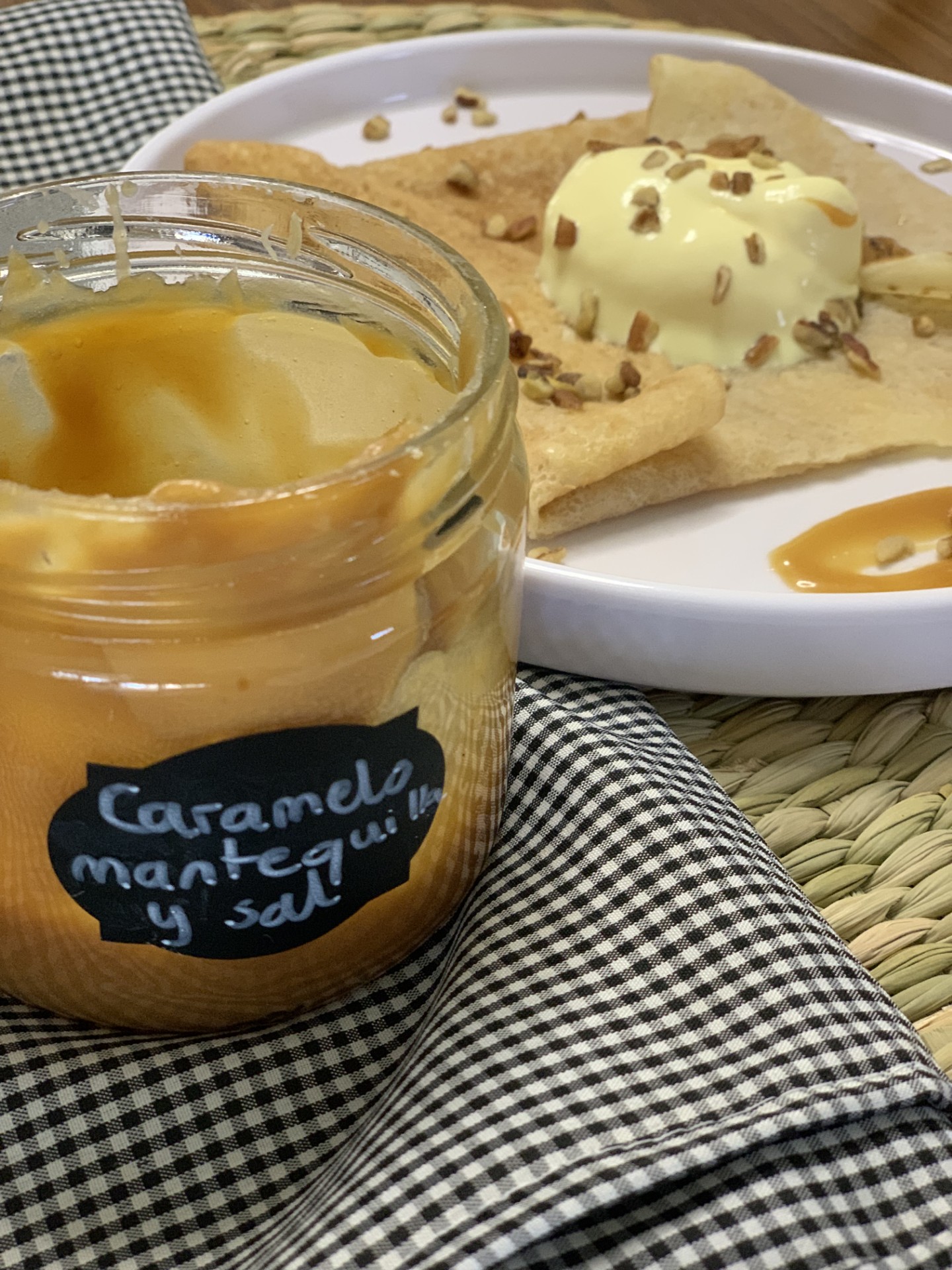 Crepes with ice cream and caramel sauce on a plate, beside a jar labeled "Caramelo mantequilla y sal" on gingham cloth.