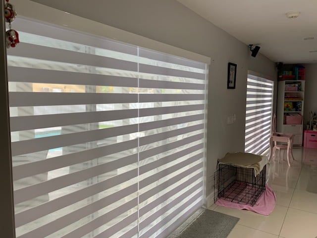 Automatic roller shades with screen material cover large windows, filtering sunlight into a living space. Motorized sunscreen blinds beige color with remote control panel on a sofa. Cityscape outside.