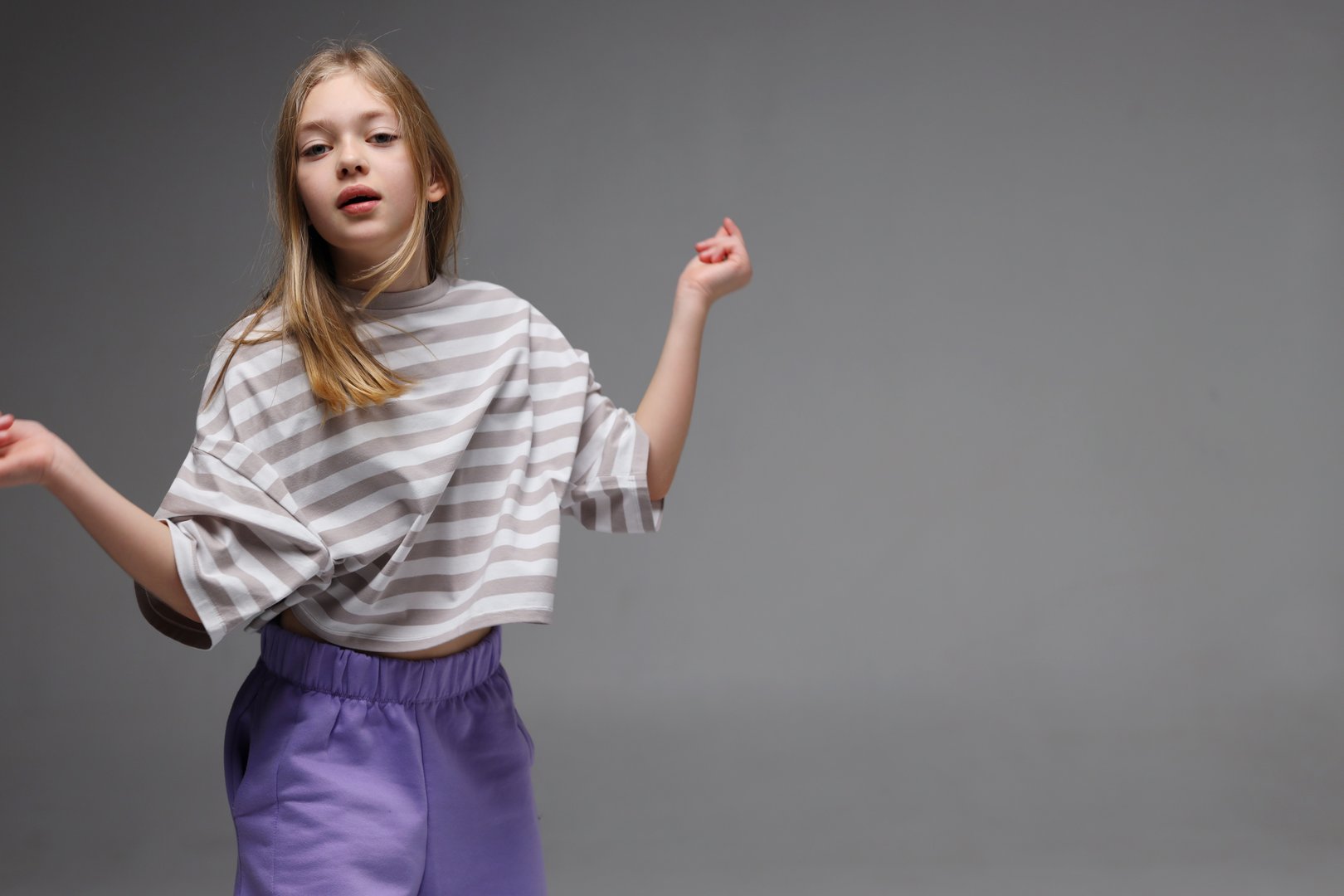Young caucasian female child posing in striped shirt and purple pants on gray background.