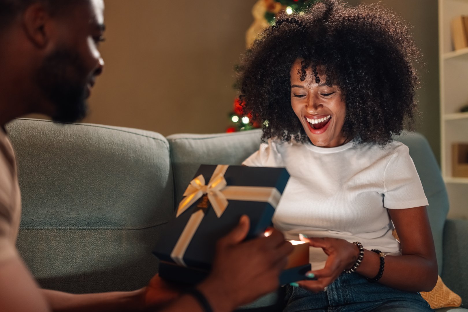 A delighted multiracial couple cherishes a tender gift exchange moment under a stunningly adorned Christmas tree, reflecting their warm connection and festive spirit in a comforting ambiance.